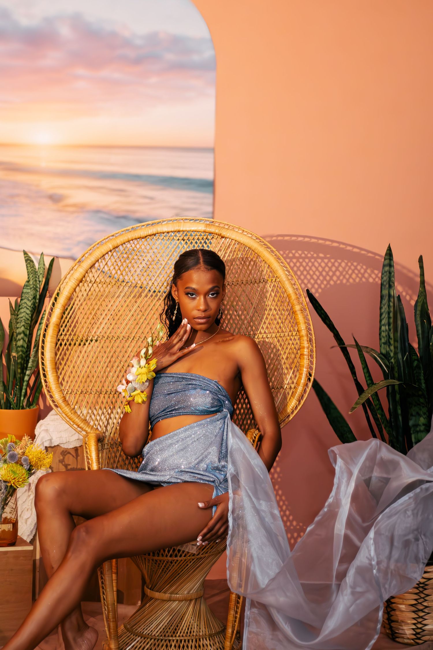 Glamour photoshoot featuring a peacock chair, flowing fabrics, and tropical plants at sunset.