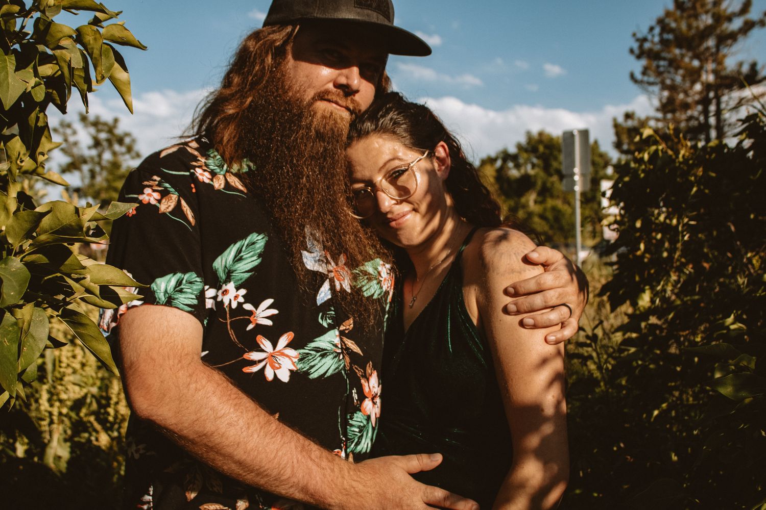 A couple embraces outdoors in warm sunlight with trees and foliage in the background.