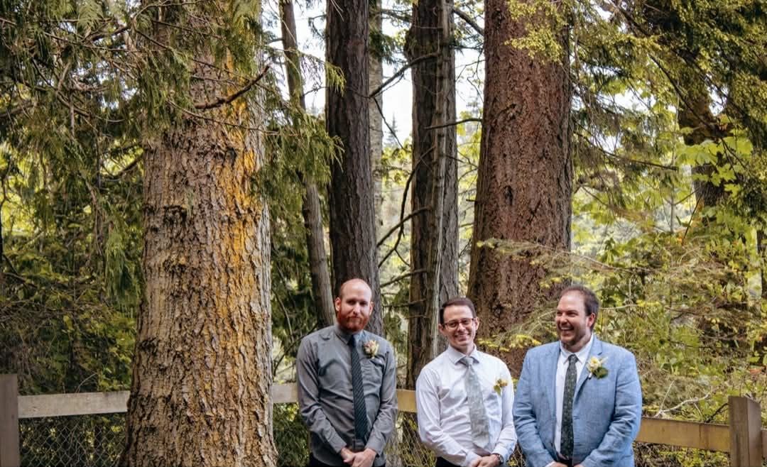 A wedding party in blue suits stands together outdoors among tall trees in a forest setting.
