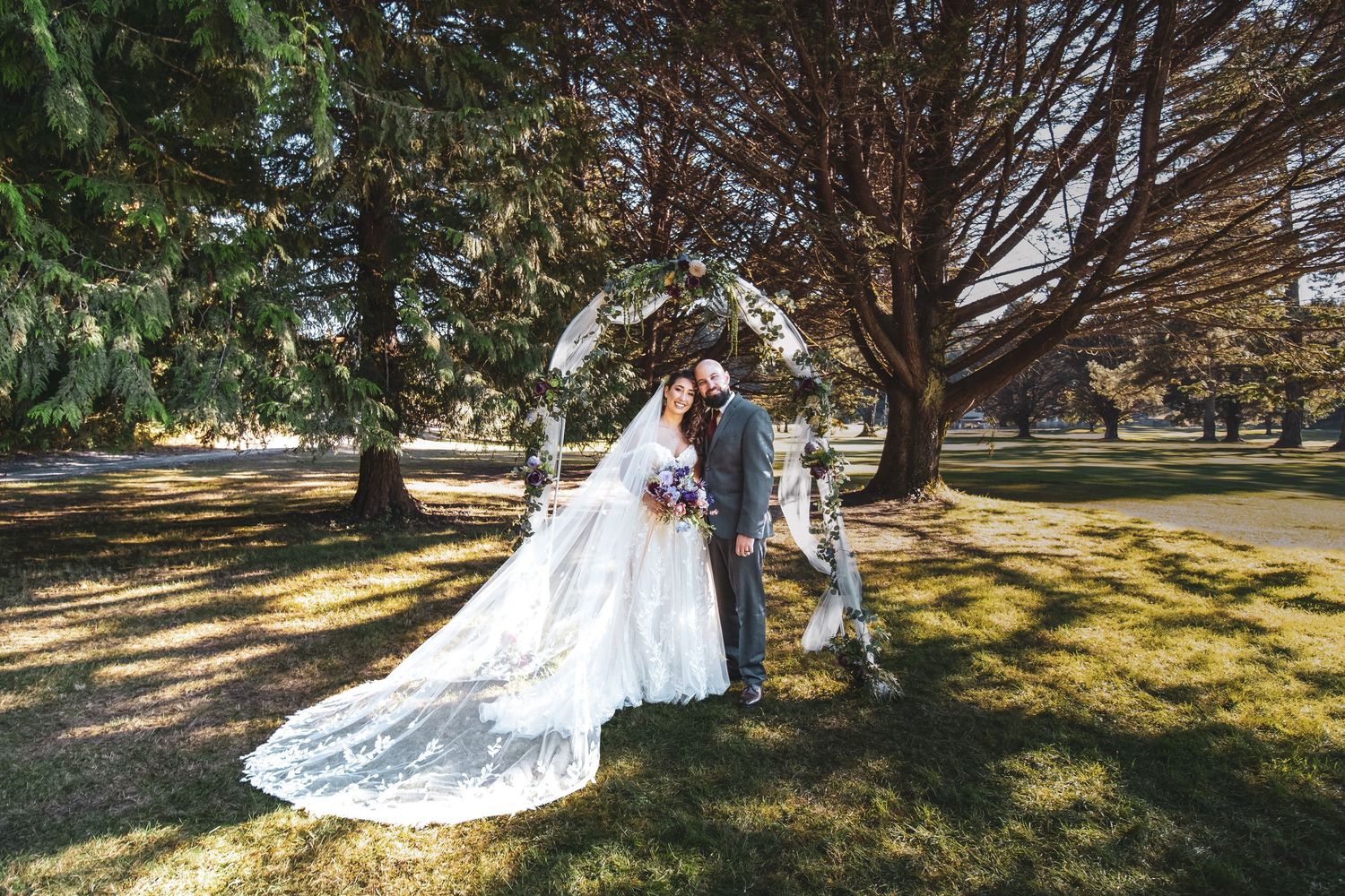 A couple shares a romantic moment under large trees in a park during their wedding, with a flowing lace veil trailing behind.