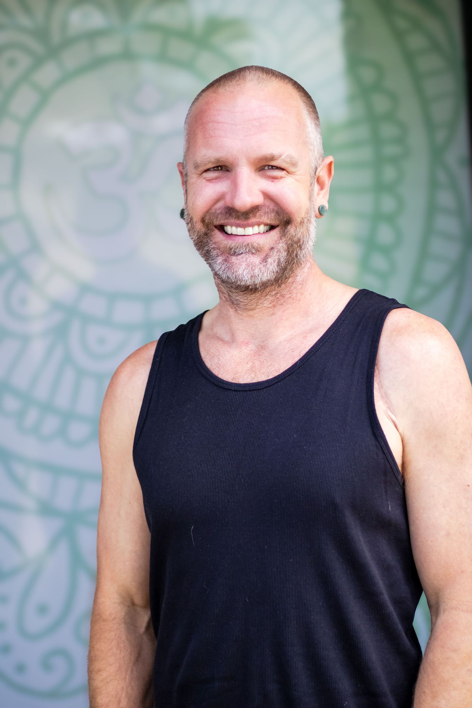 A person in a black tank top smiles warmly against a decorative green patterned background.