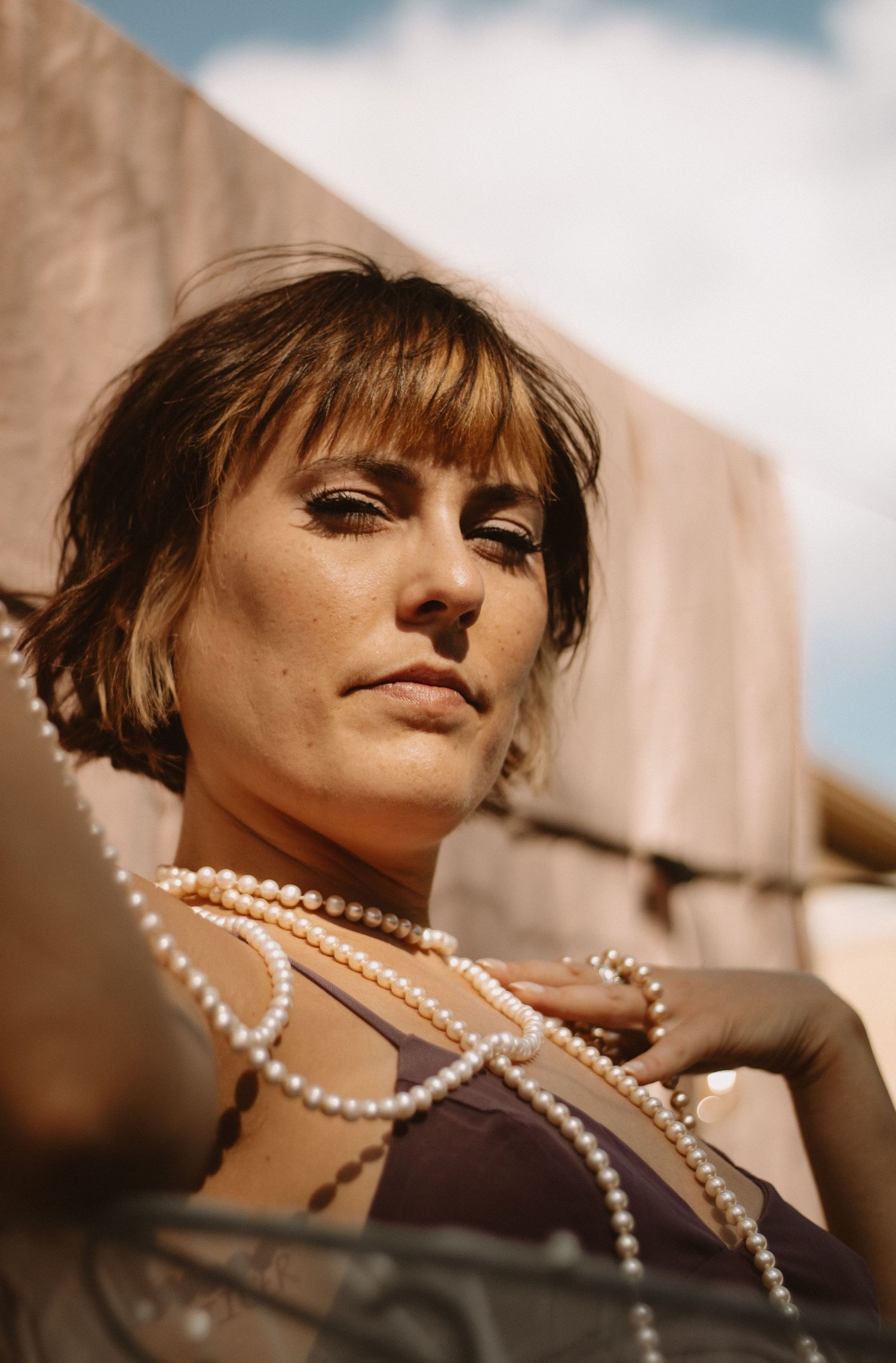 A fashion portrait shows a model wearing a pearl necklace in warm sunlight against a beige wall.