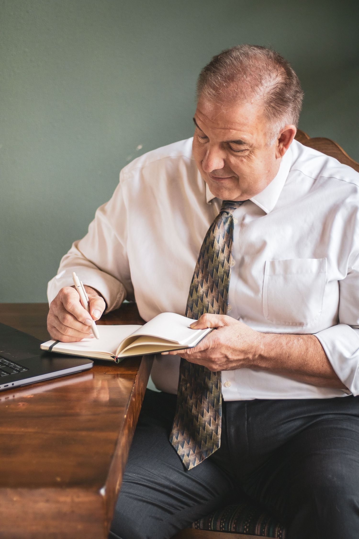 A person in a white shirt and tie leans over a wooden desk while writing in a small notebook.