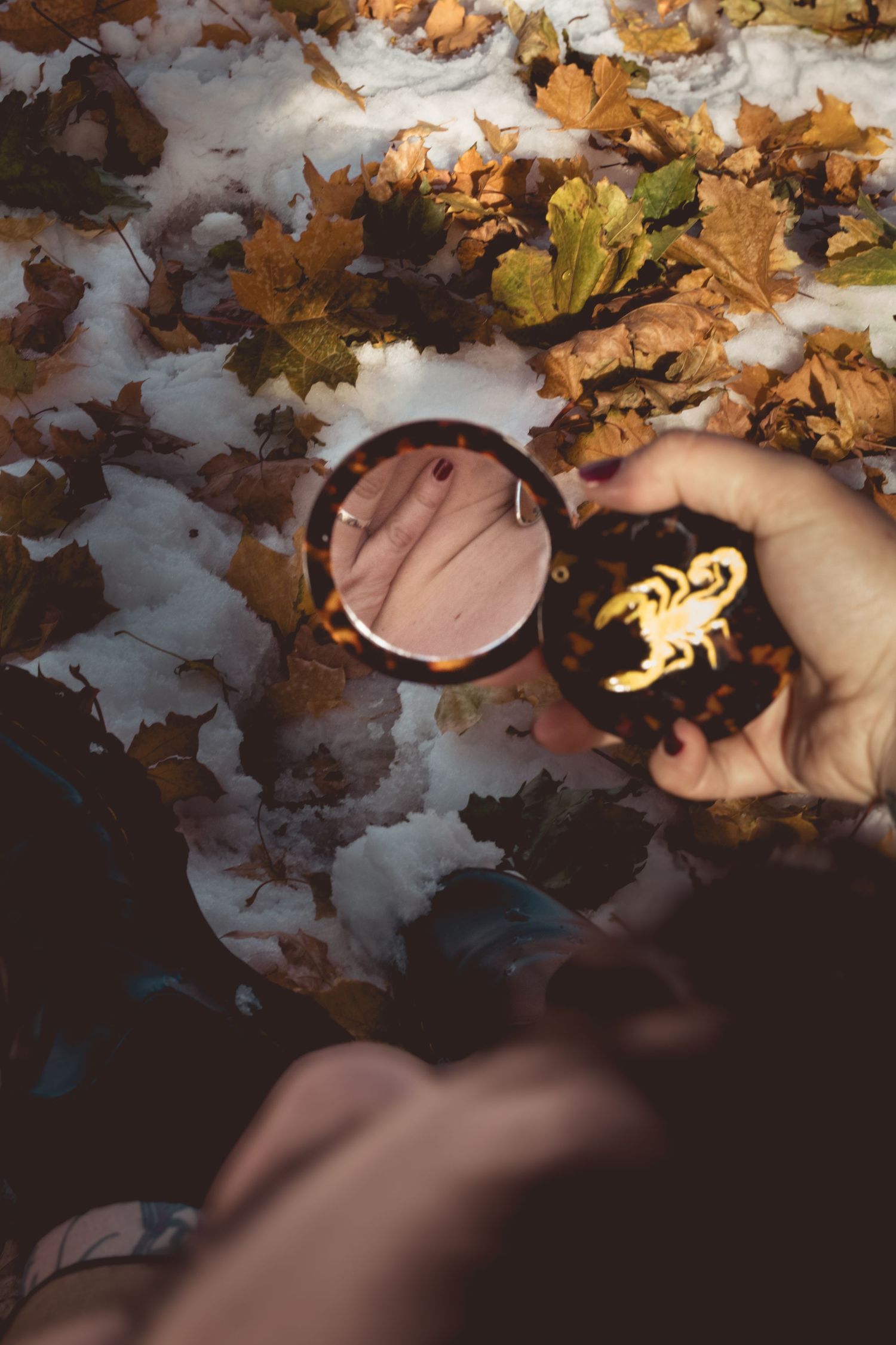 A hand holds a lit match among scattered autumn leaves and snow on the ground in moody lighting.