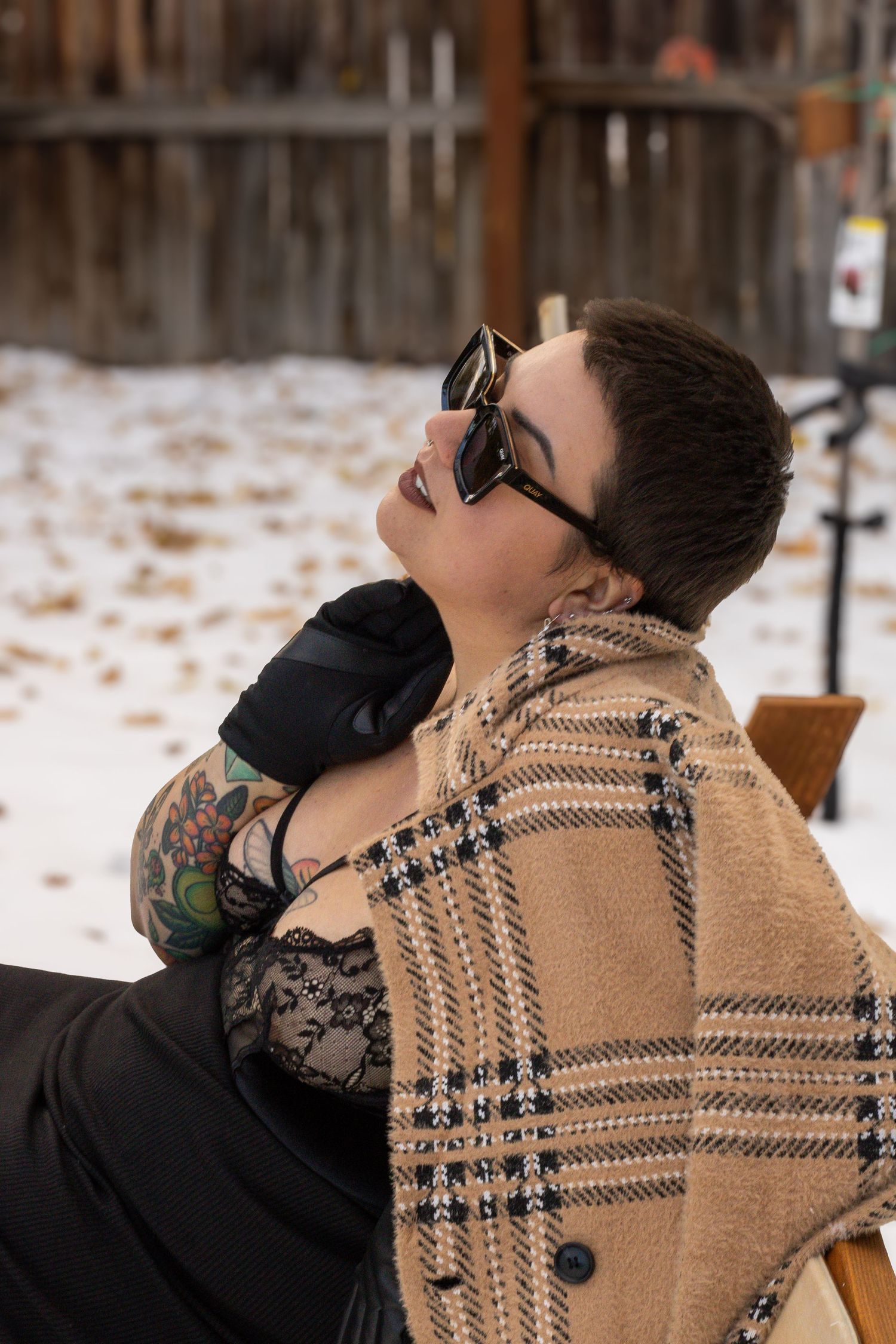 Someone wearing sunglasses and a beige plaid coat reclines outdoors in winter against a snowy backdrop.