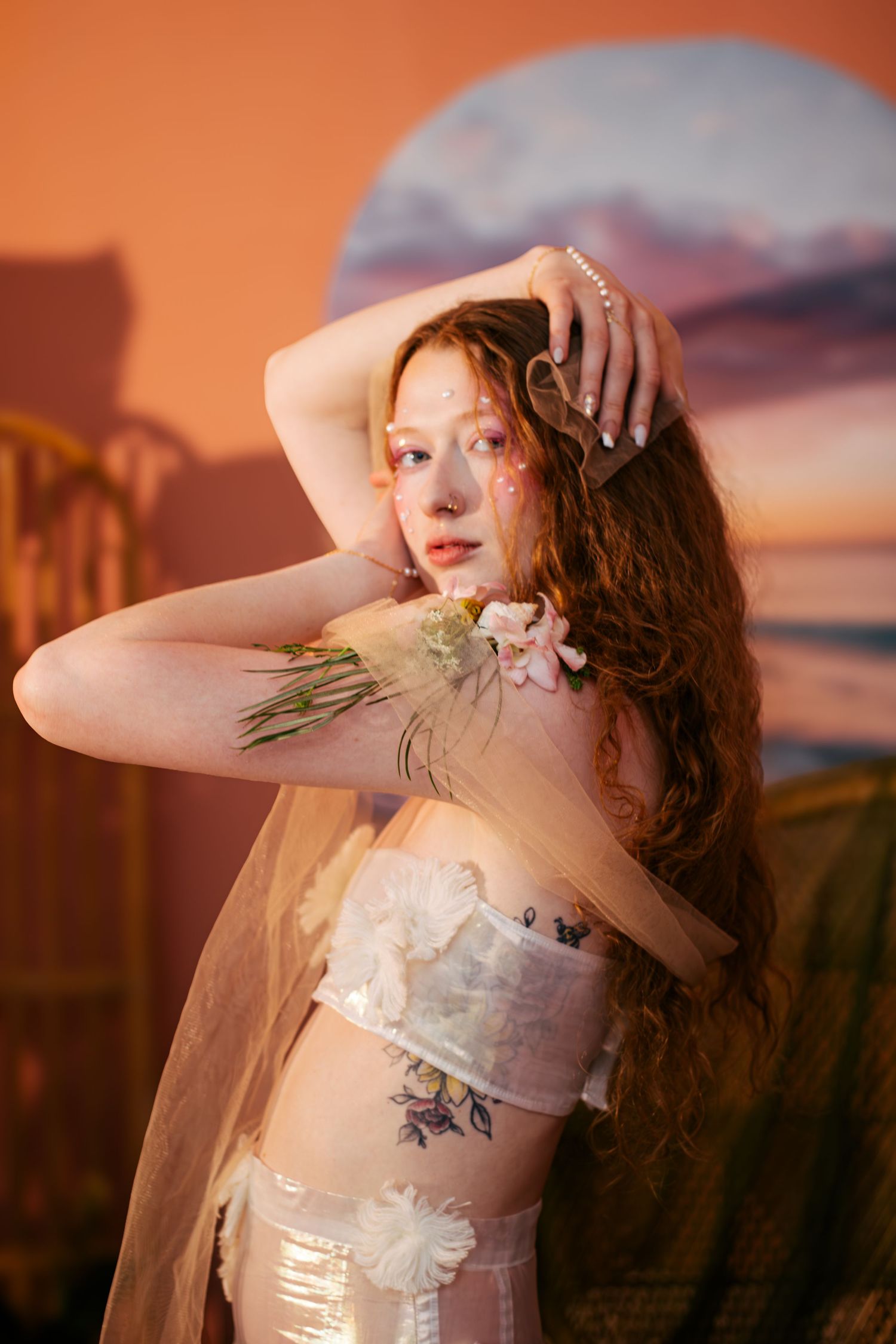 Ethereal portrait in soft lighting with delicate floral details and sheer fabric against an orange-tinted sunset backdrop.