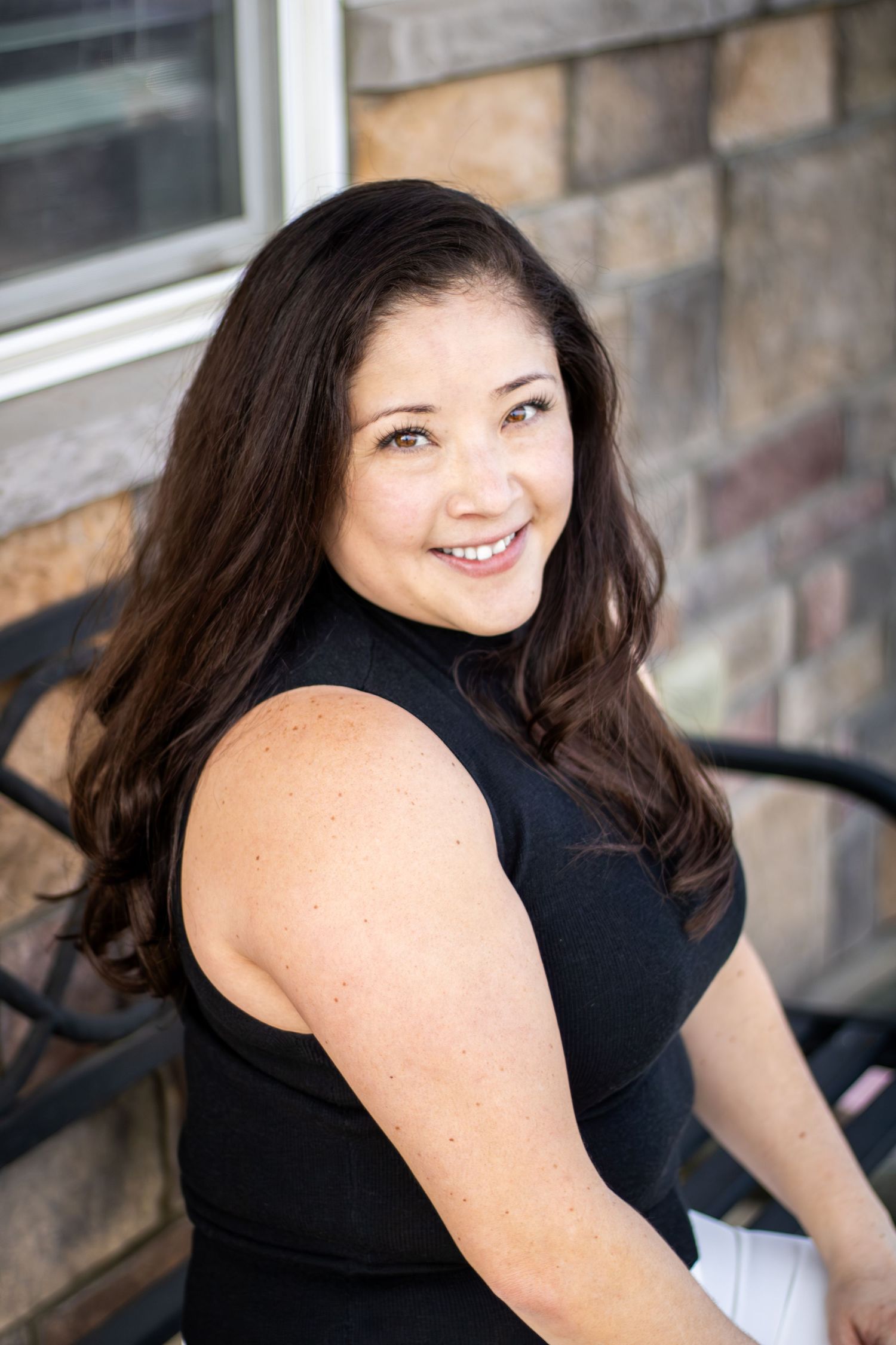 Casual outdoor portrait of someone in dark sleeveless attire smiling while seated against a brick exterior.