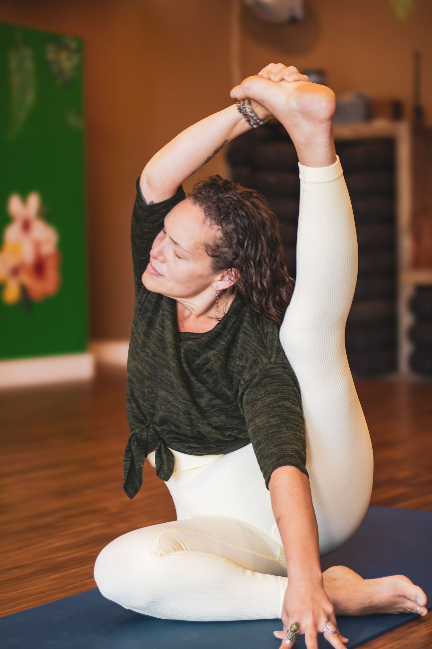 Person in olive green top and white leggings performs an advanced yoga pose on a blue mat in a studio setting.