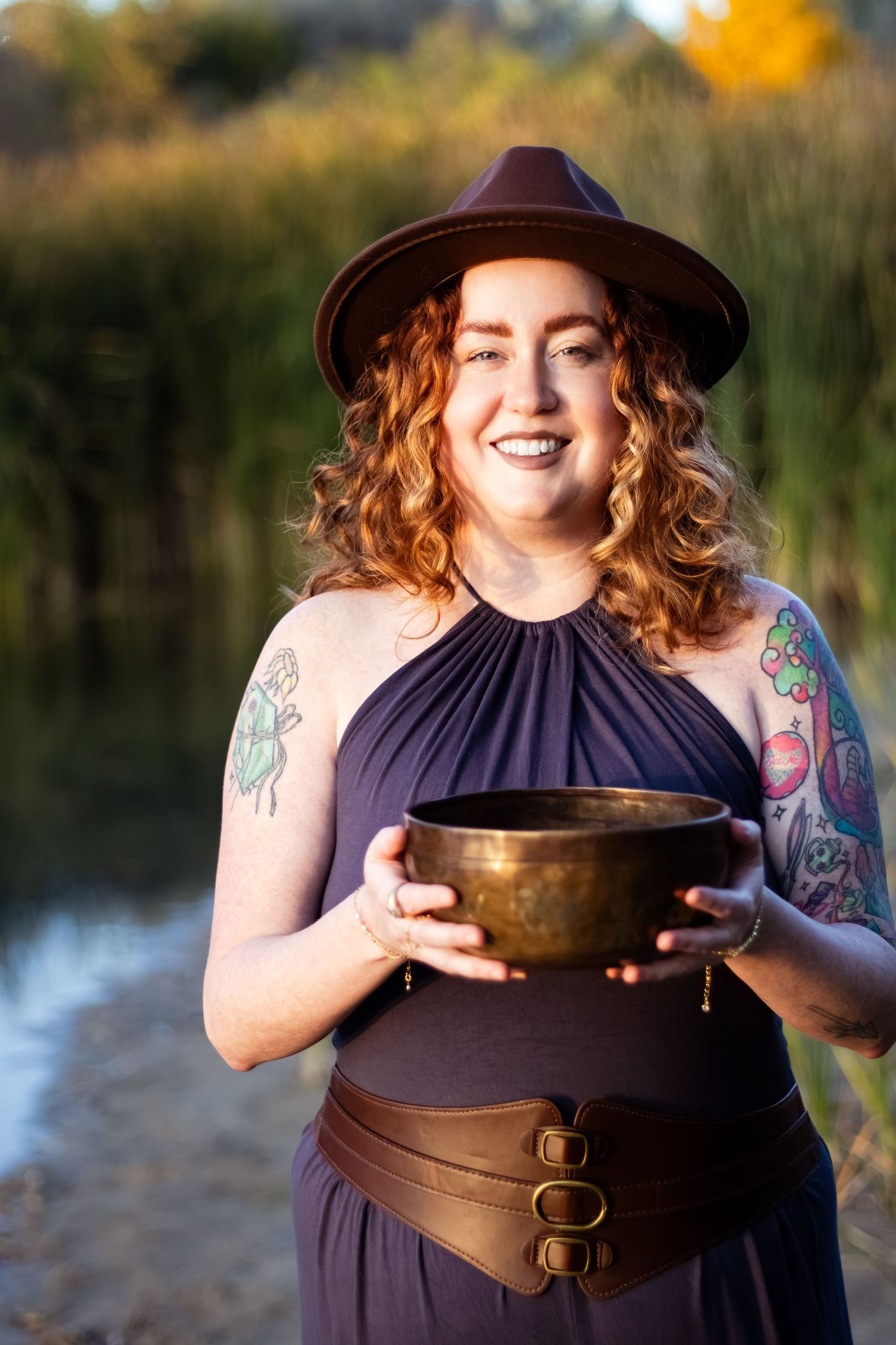 A person holds a meditation bowl while wearing a navy dress with brown leather belt outdoors at sunset.