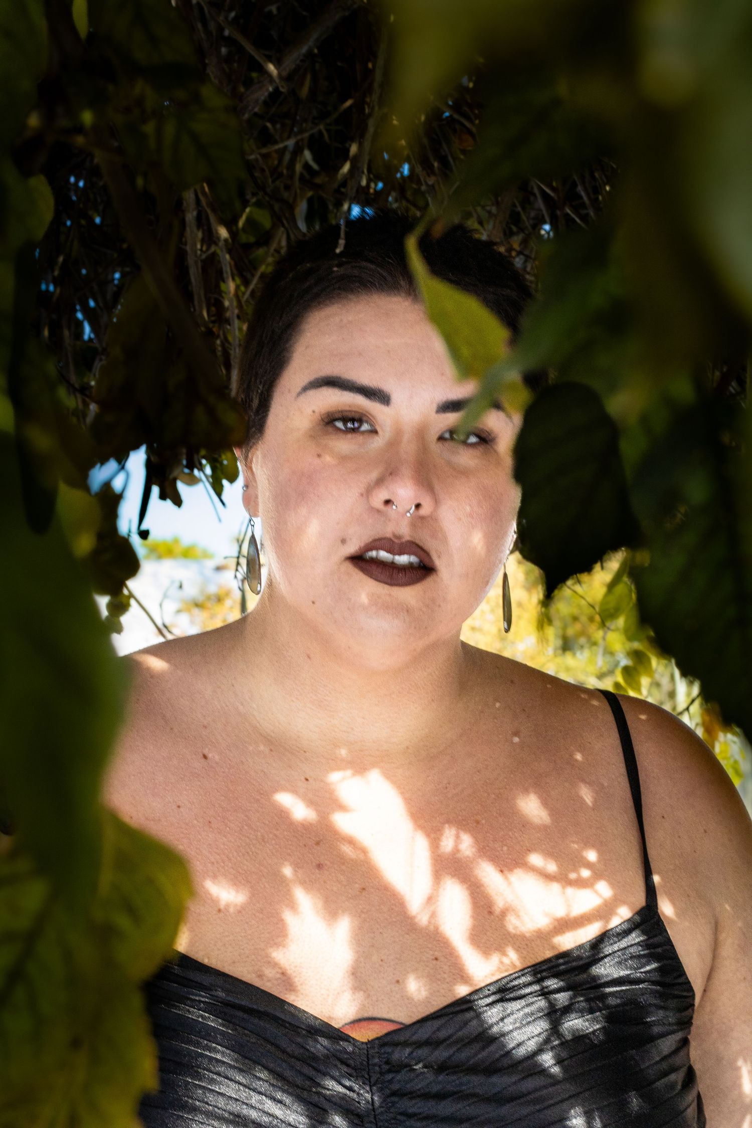 Portrait in dappled sunlight wearing black satin against lush green foliage background.