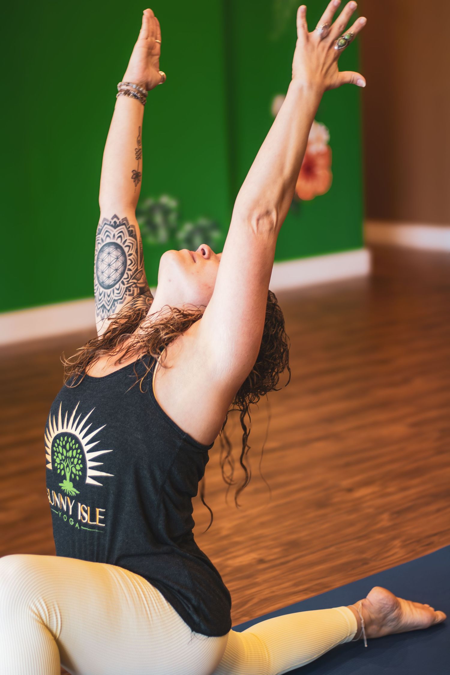 A person practicing yoga on a wooden floor wearing a black tank top with a sun design performs a stretching pose.