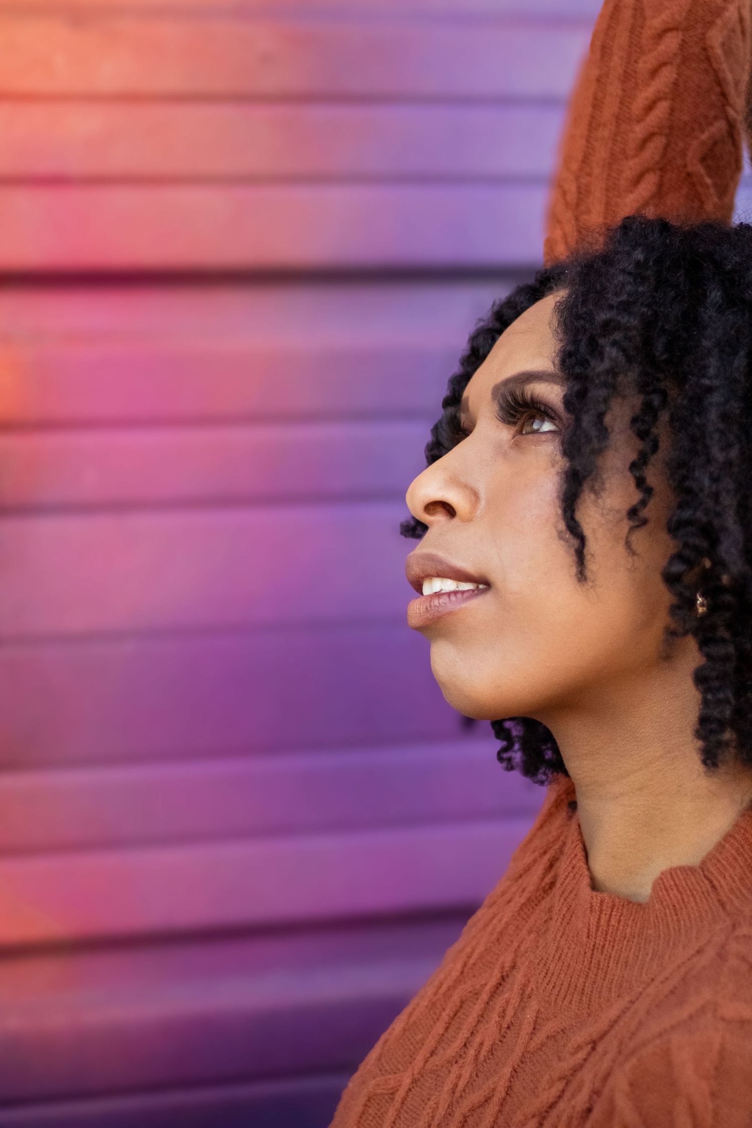 Side profile portrait against purple wall wearing brown sweater with natural curly hair and looking upward.