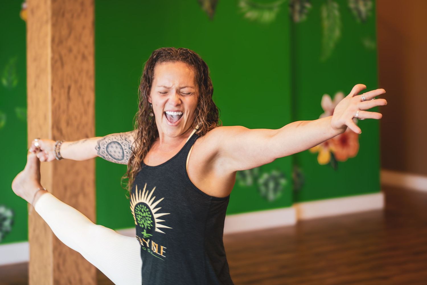 A person wearing a black tank top stretches into a yoga pose with outstretched arms against a green wall.