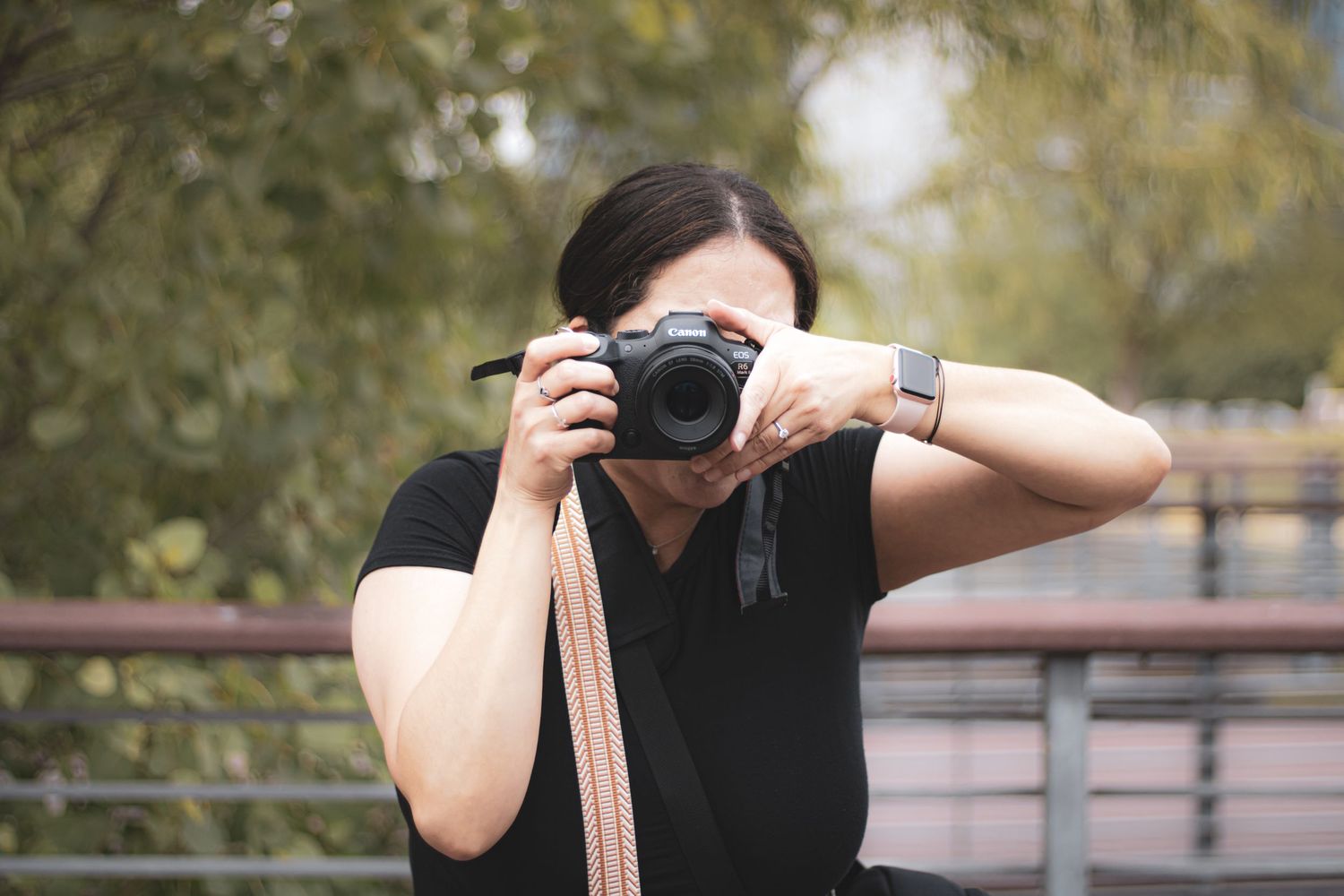 A photographer wearing a black shirt holds up a digital camera with a pink camera strap outdoors.