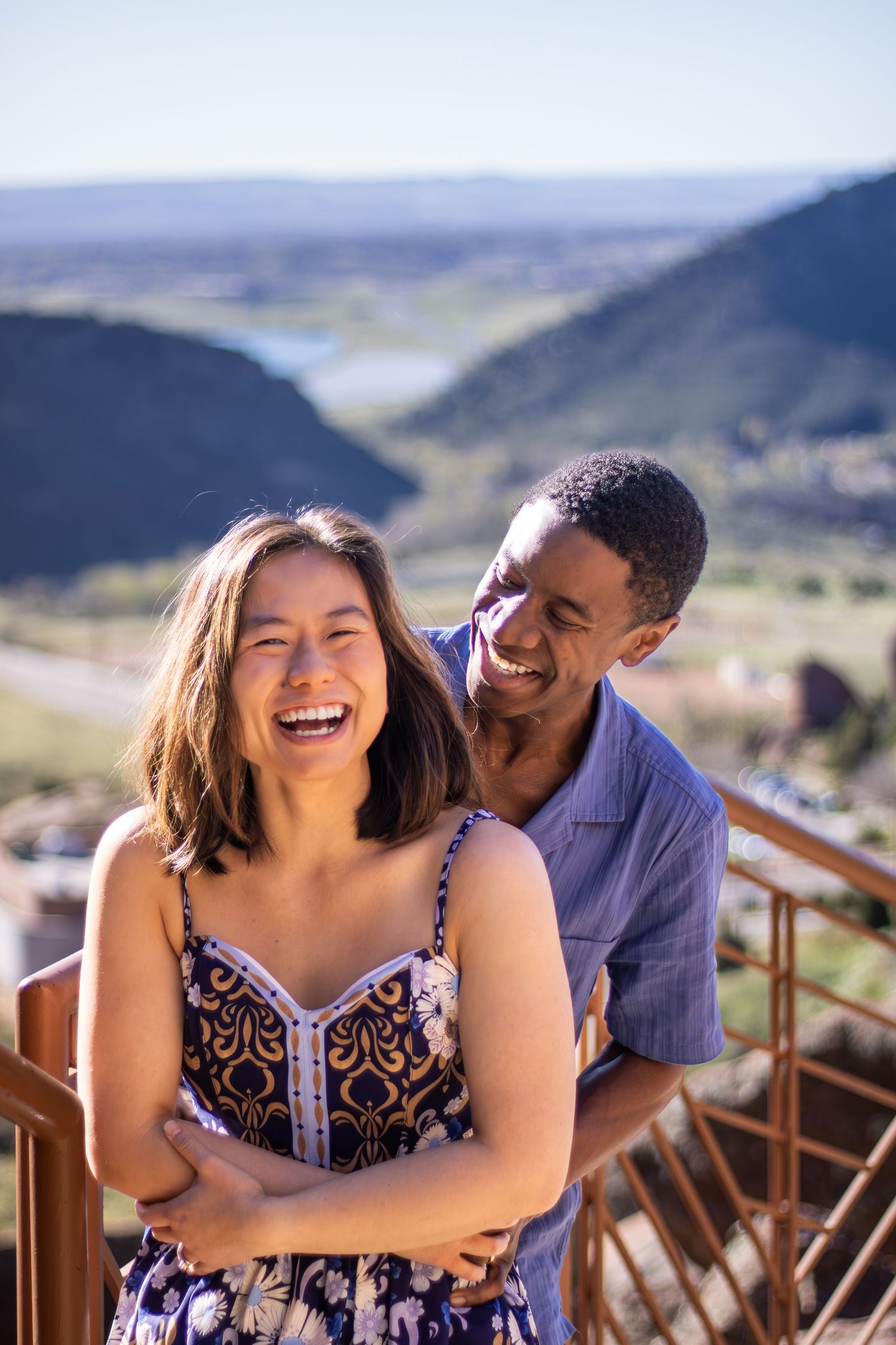 A romantic couple embraces and laughs together on a deck overlooking a mountainous landscape.