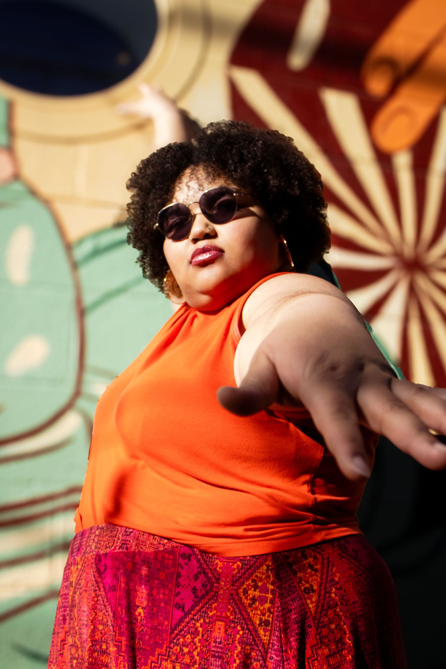 Person in orange tank top and red patterned skirt dancing against a retro-styled wall with starburst pattern.
