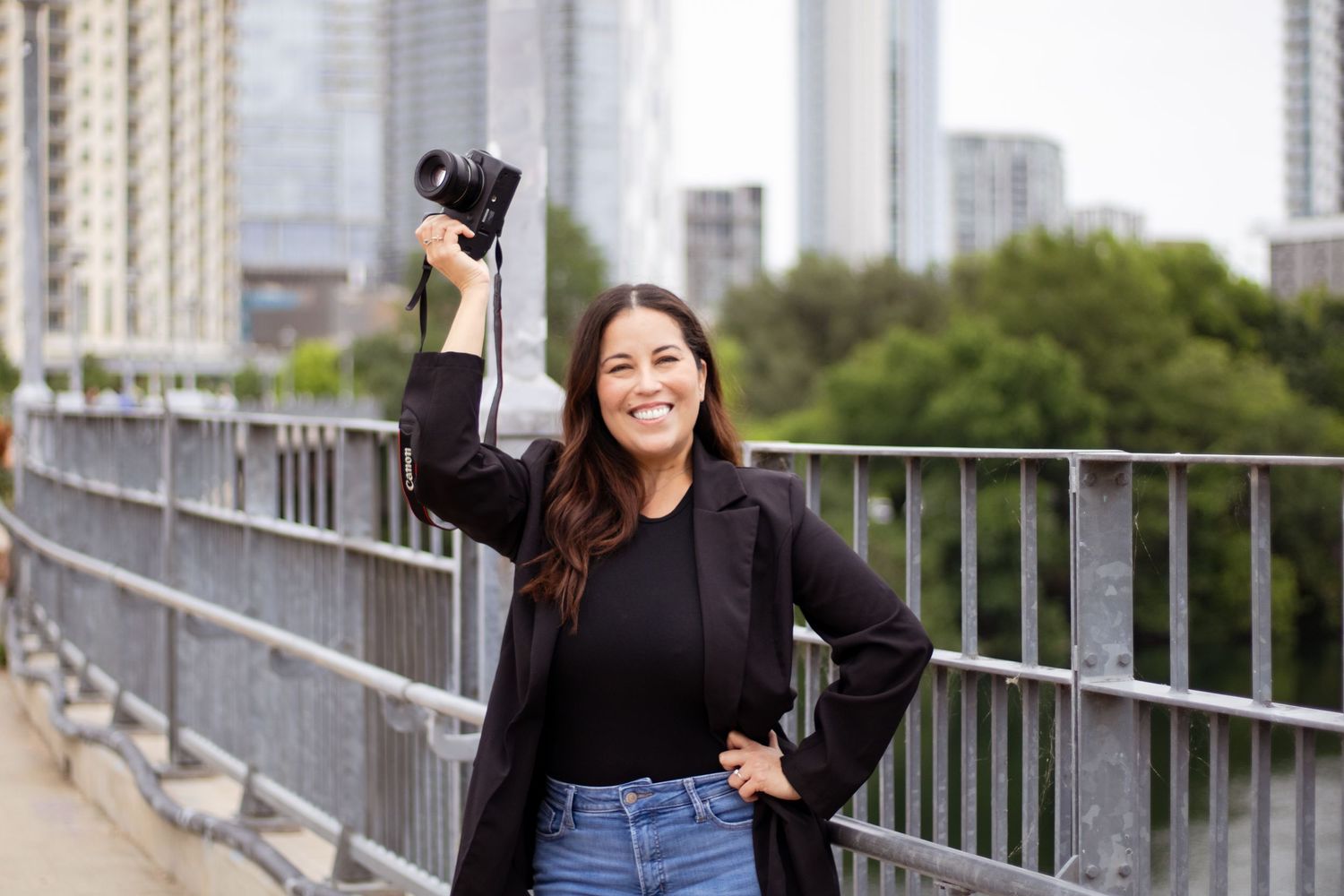 A person in a black jacket and jeans poses on a bridge walkway with city buildings visible in the background.