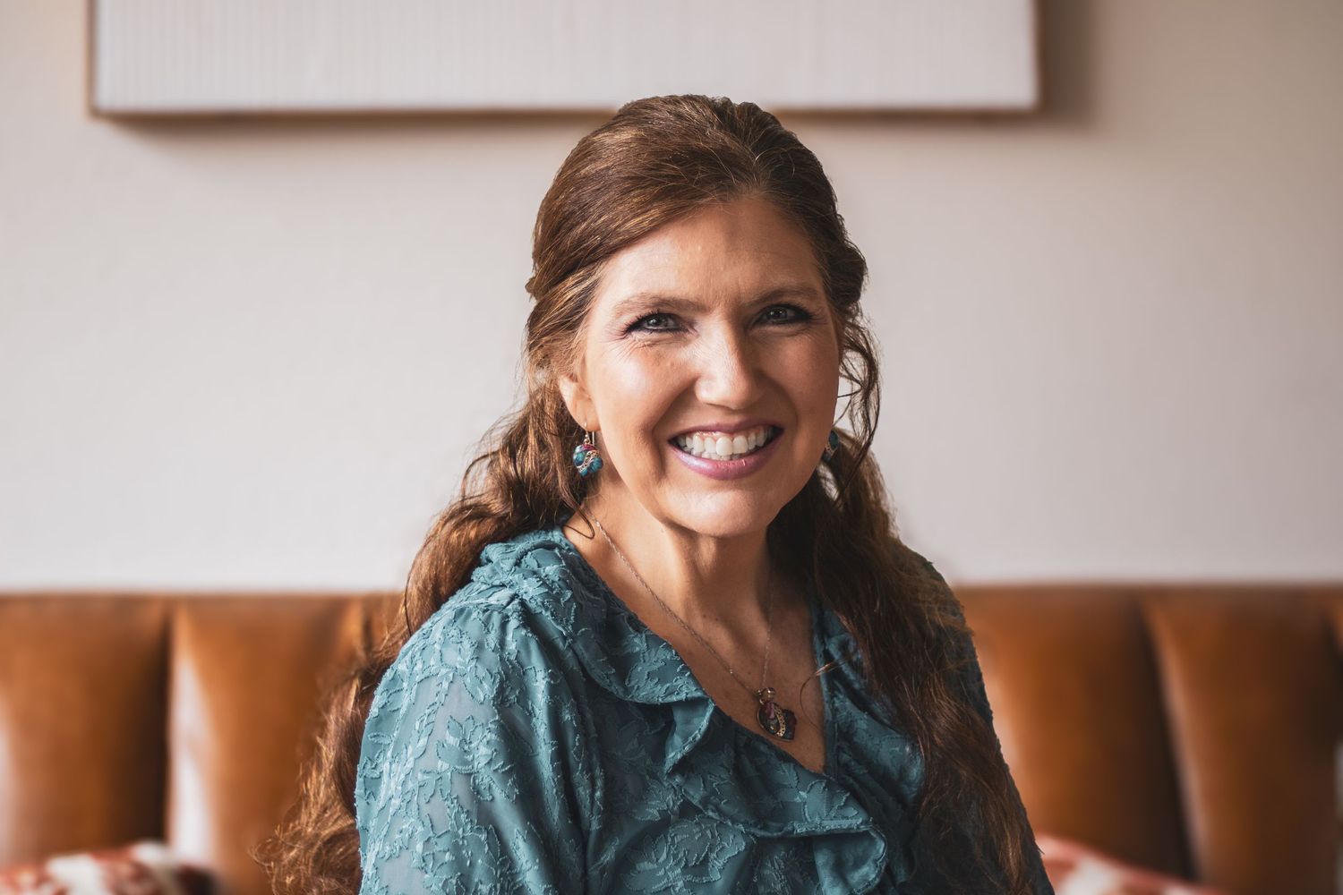 A woman in a teal dress sits on a brown leather couch while smiling warmly at the camera.