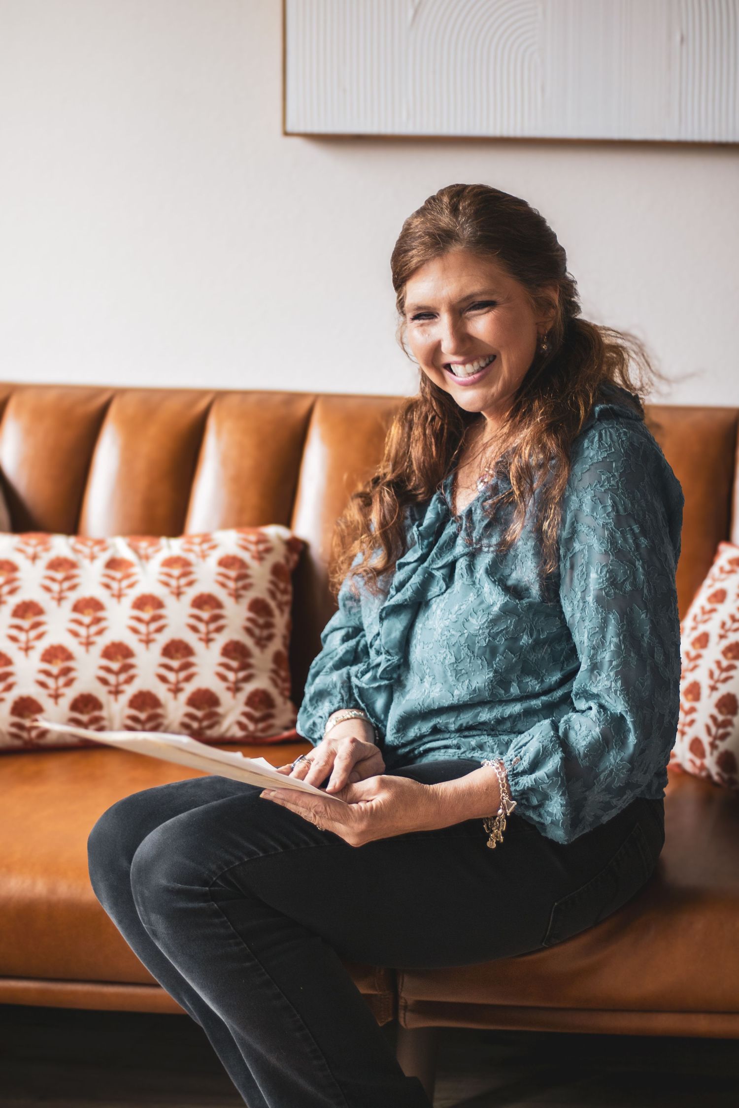 Person in teal blouse relaxes on brown leather couch while looking at phone and smiling.