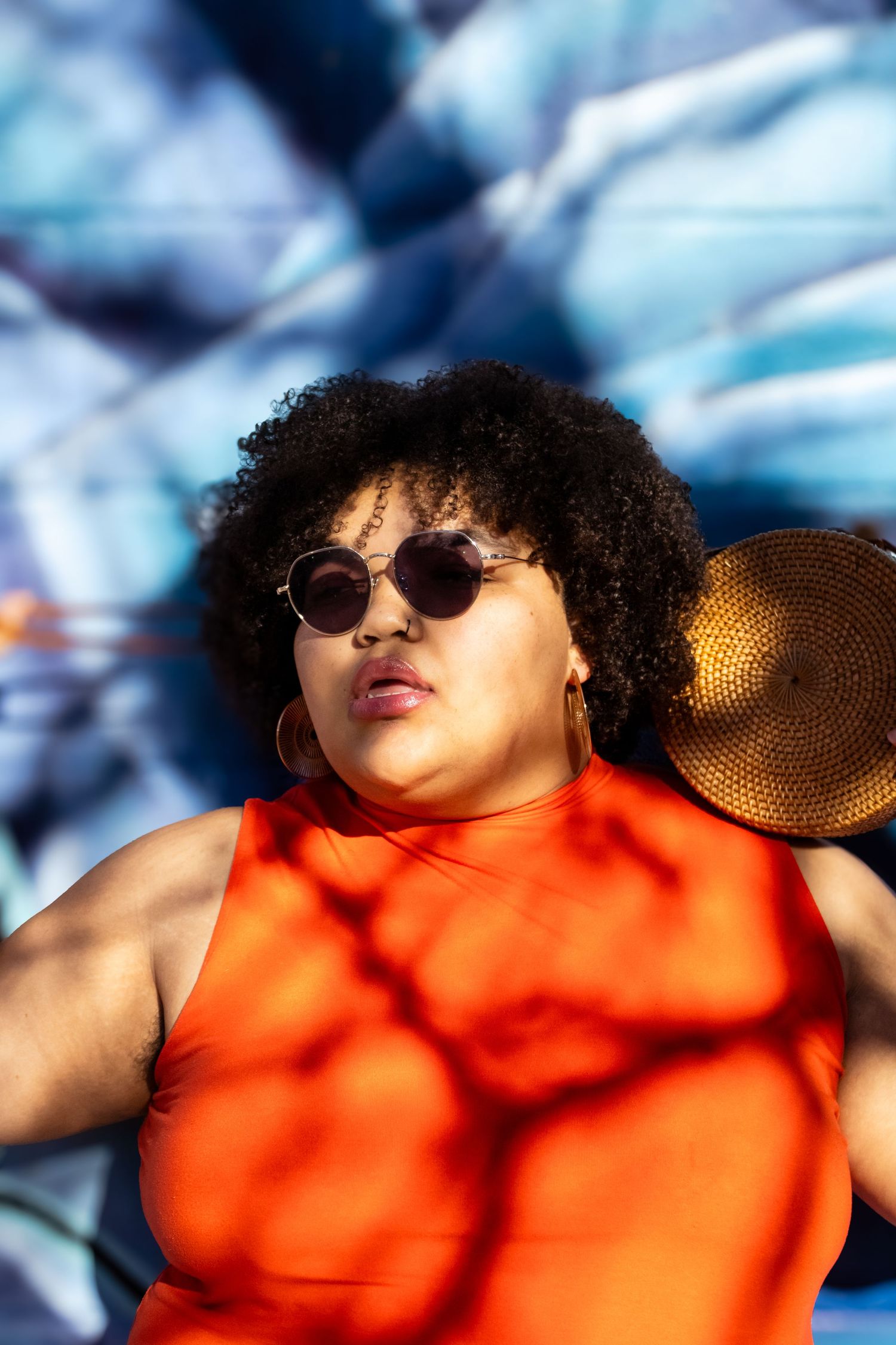 Person in orange sleeveless top and sunglasses relaxing outdoors on a sunny day against blue background.