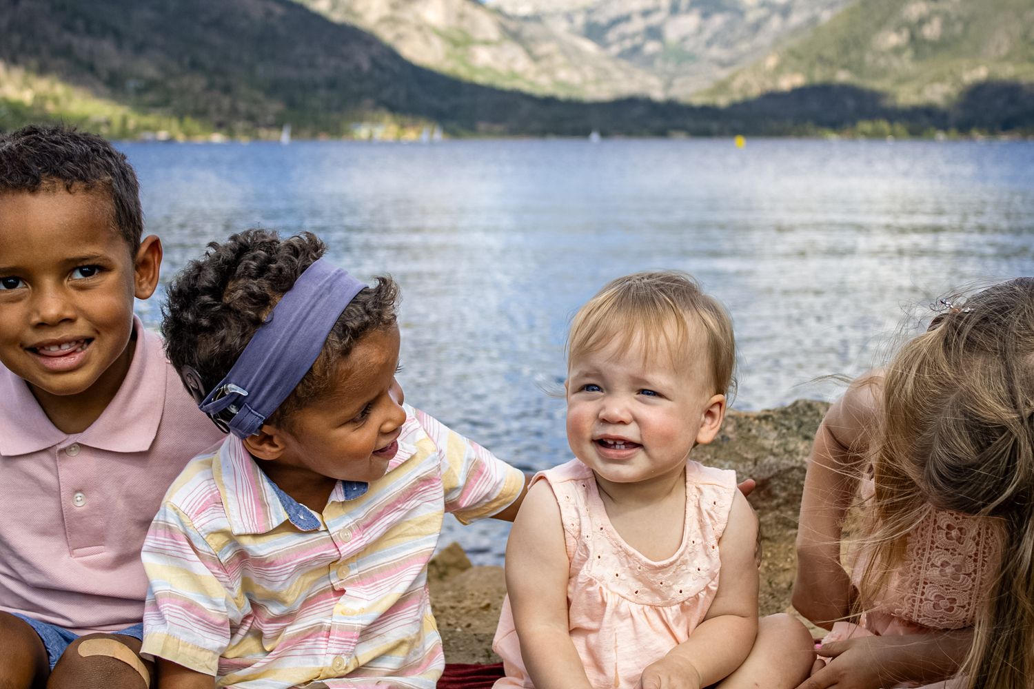 Children sit together by a lake on a sunny day with mountains visible in the distance.