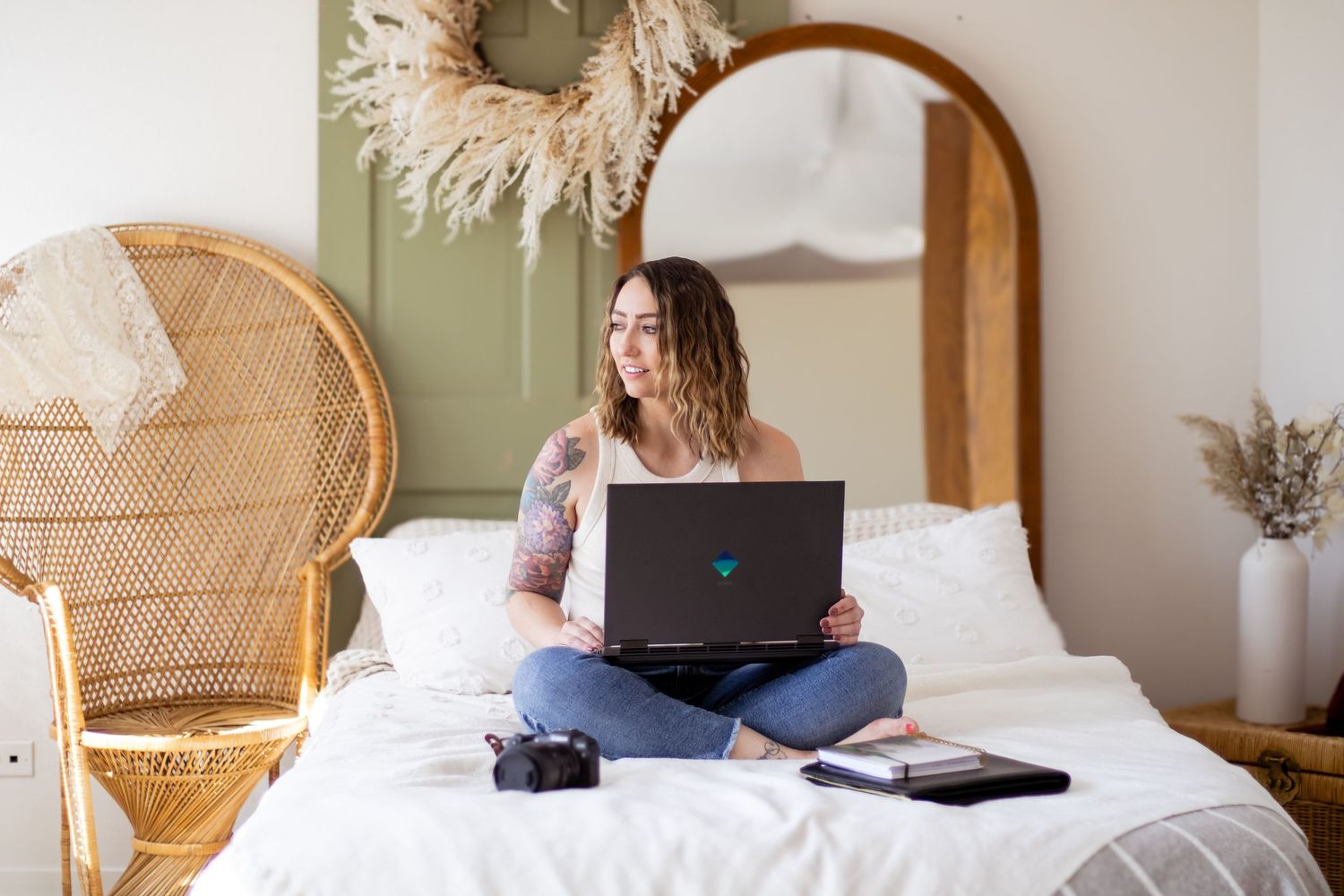 Person sitting cross-legged on white bedding while working on laptop in cozy bedroom with wicker decor.