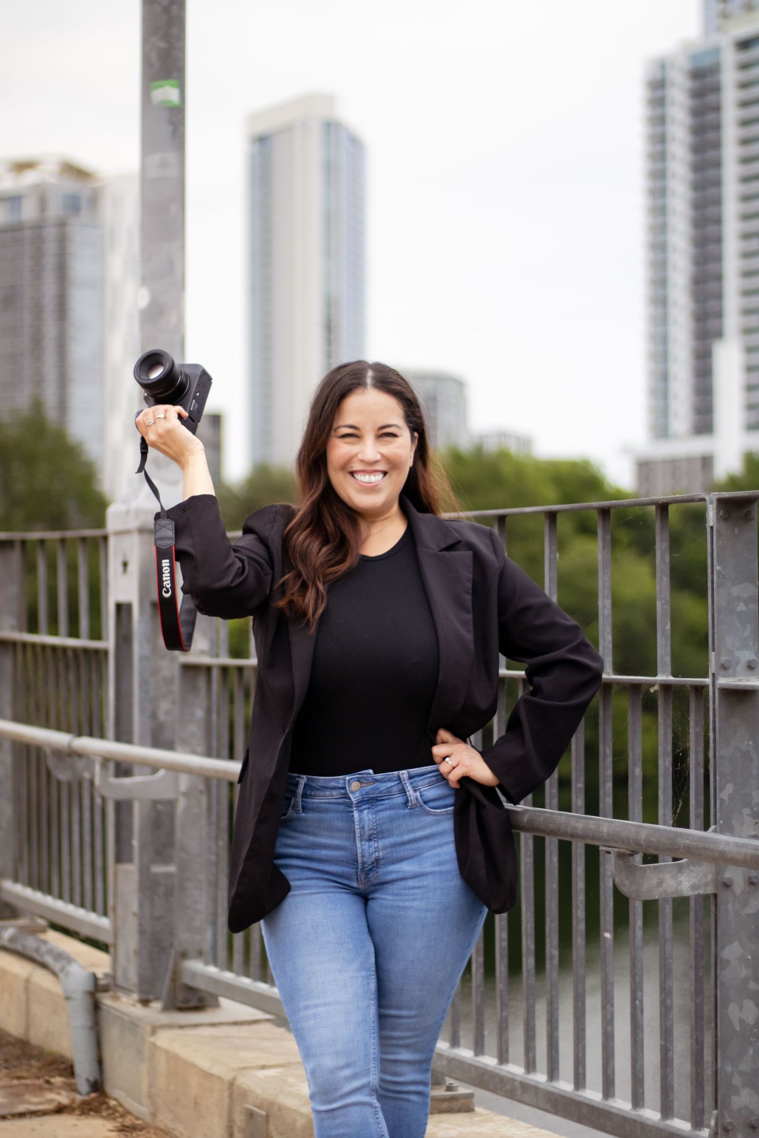 A photographer in a black blazer and jeans holds up a camera while posing on an urban bridge with city buildings behind.