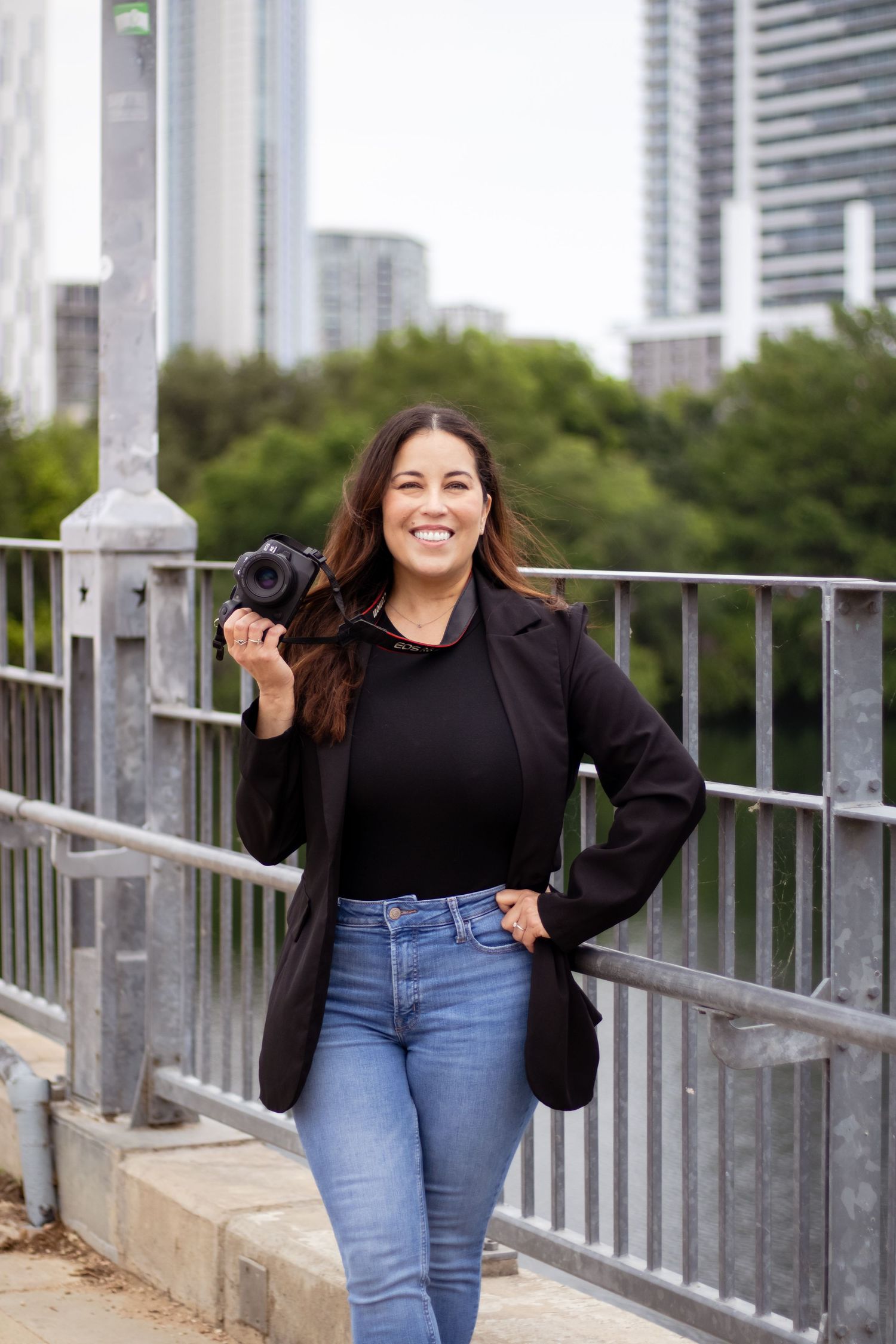 A woman in a black sweater and blue jeans poses confidently along an urban railing with city buildings in the background.