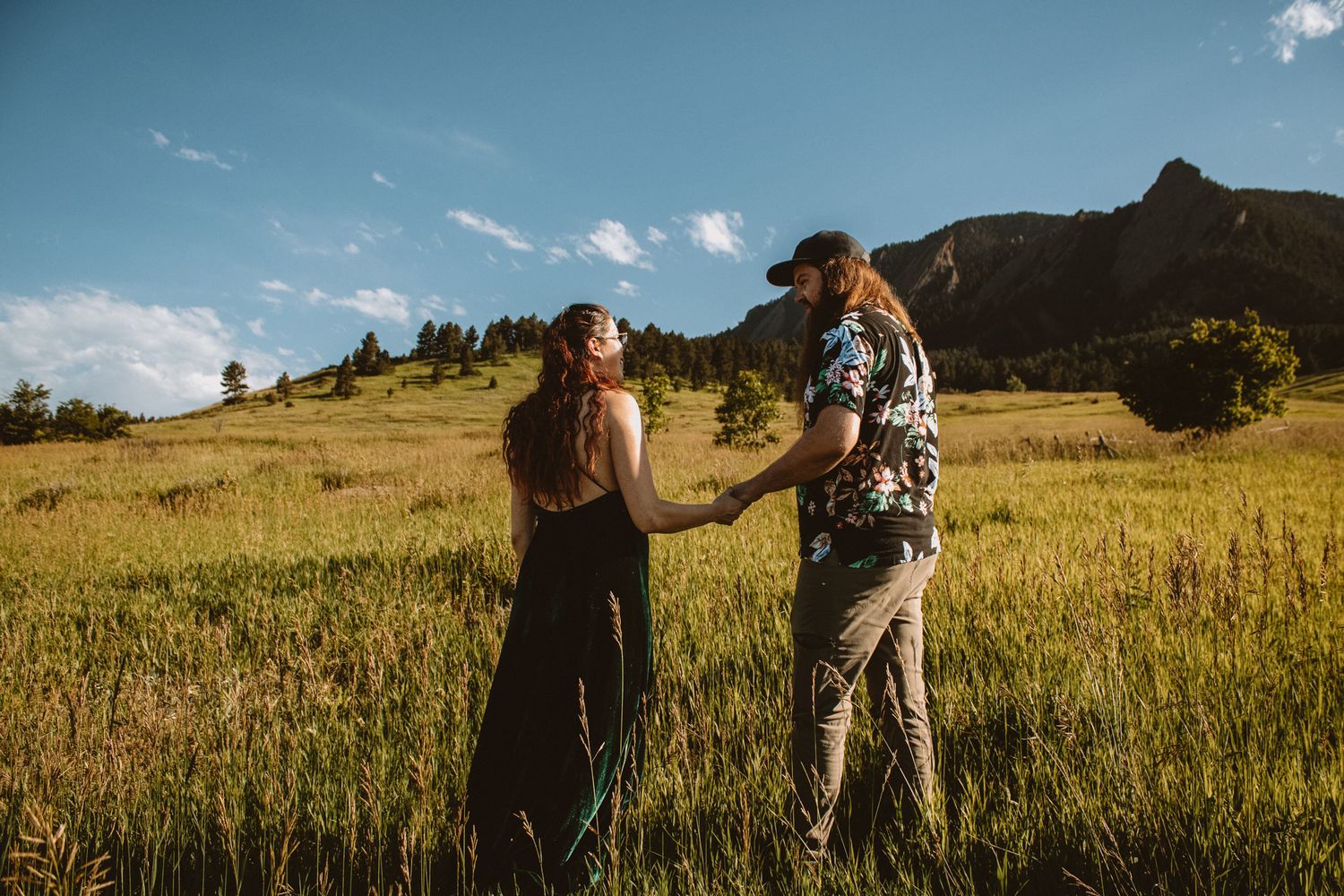 Two people walk through a golden meadow at sunset with mountains in the background.