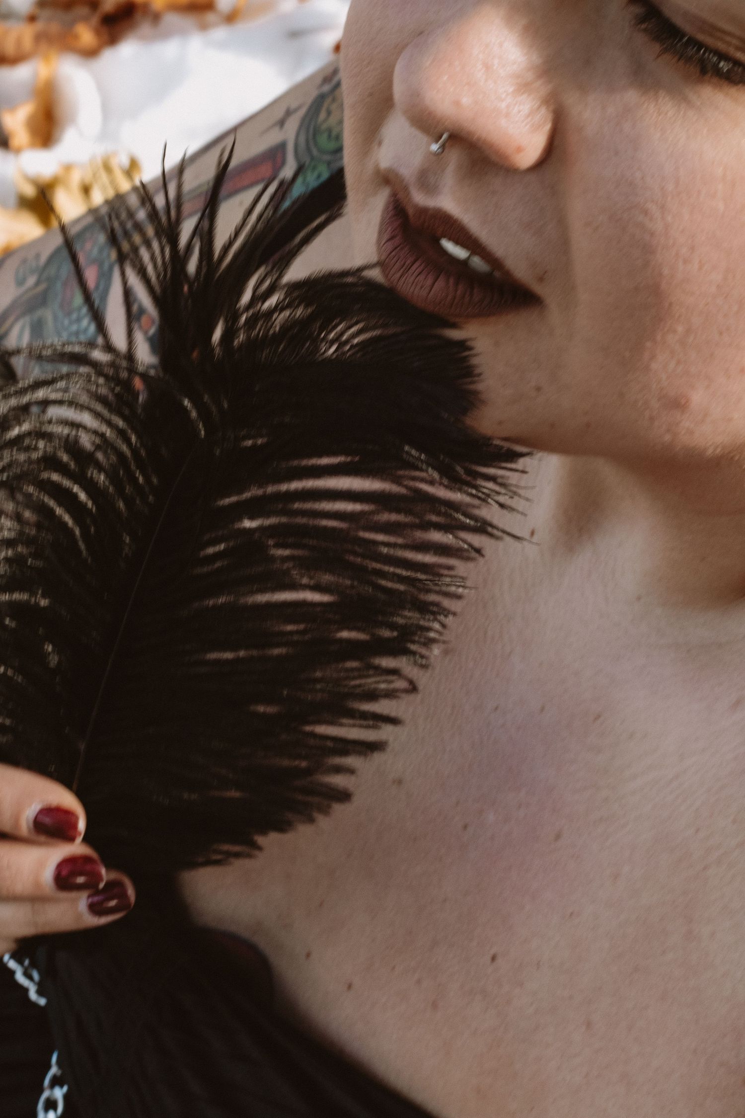 A close-up shot shows dark red lips and nails holding a black feather against pale skin in a glamorous style.