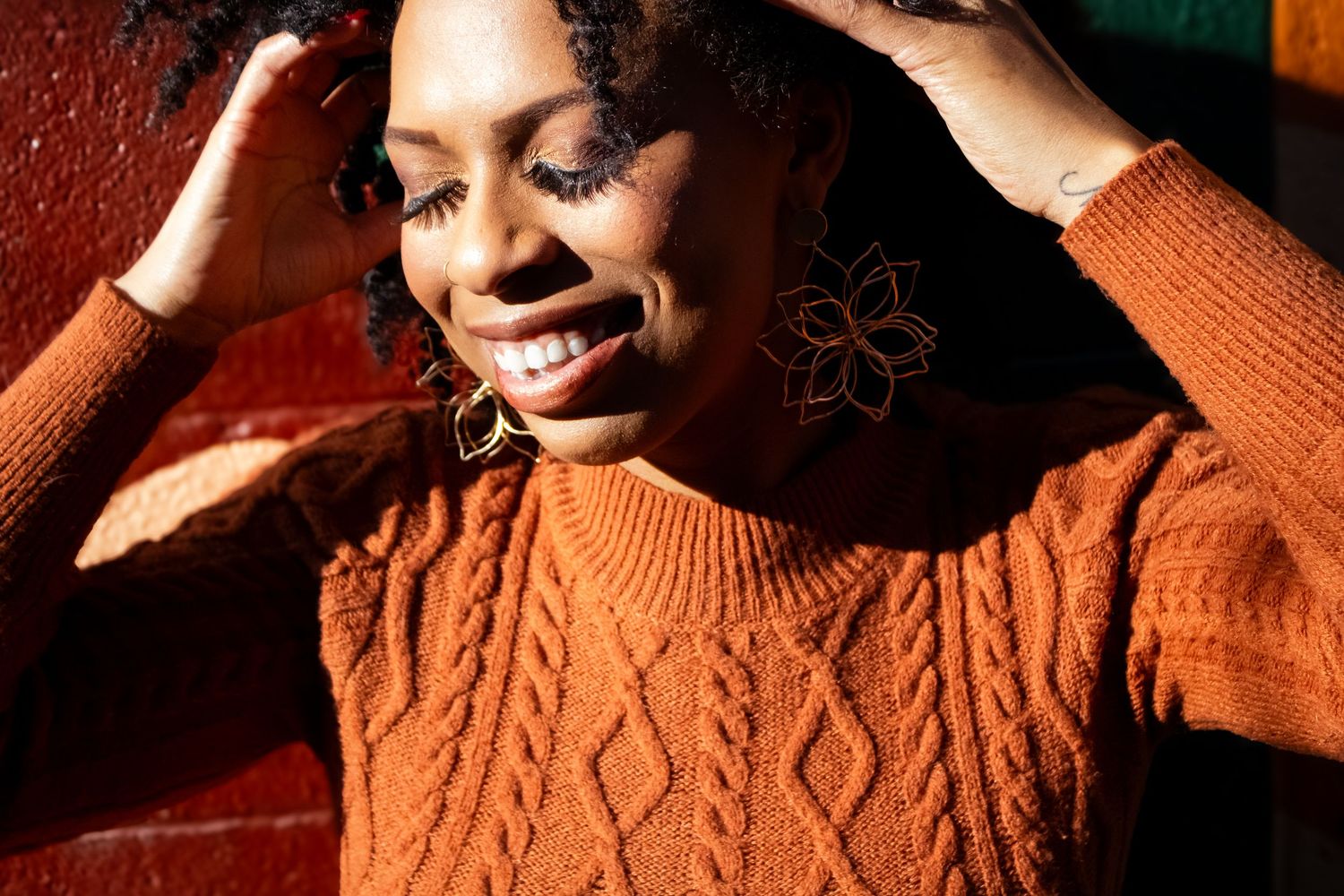 Woman in orange sweater poses expressively in warm lighting against dark background.