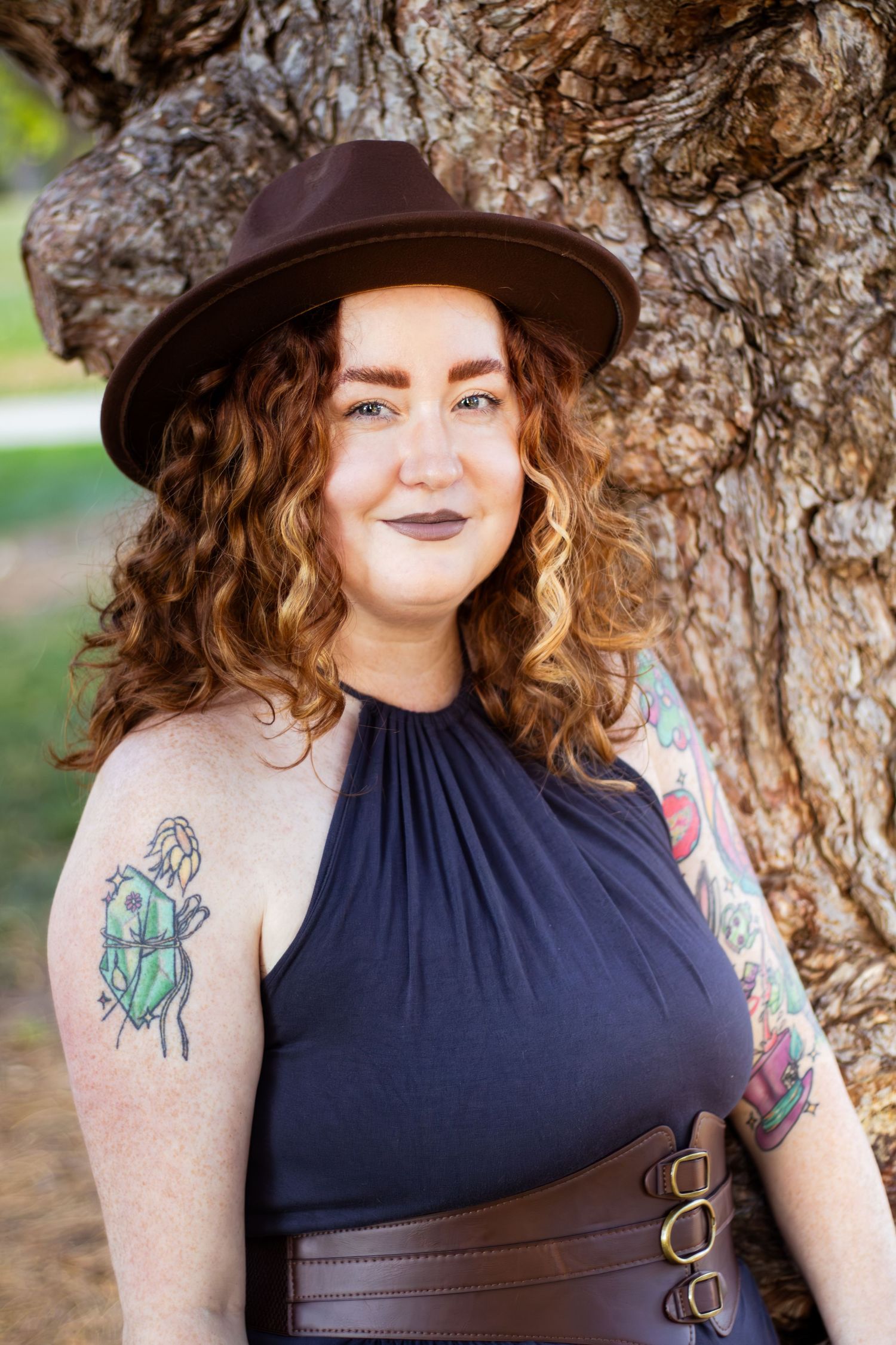 Person in a navy halter top and brown hat poses against a textured tree trunk in an outdoor setting.