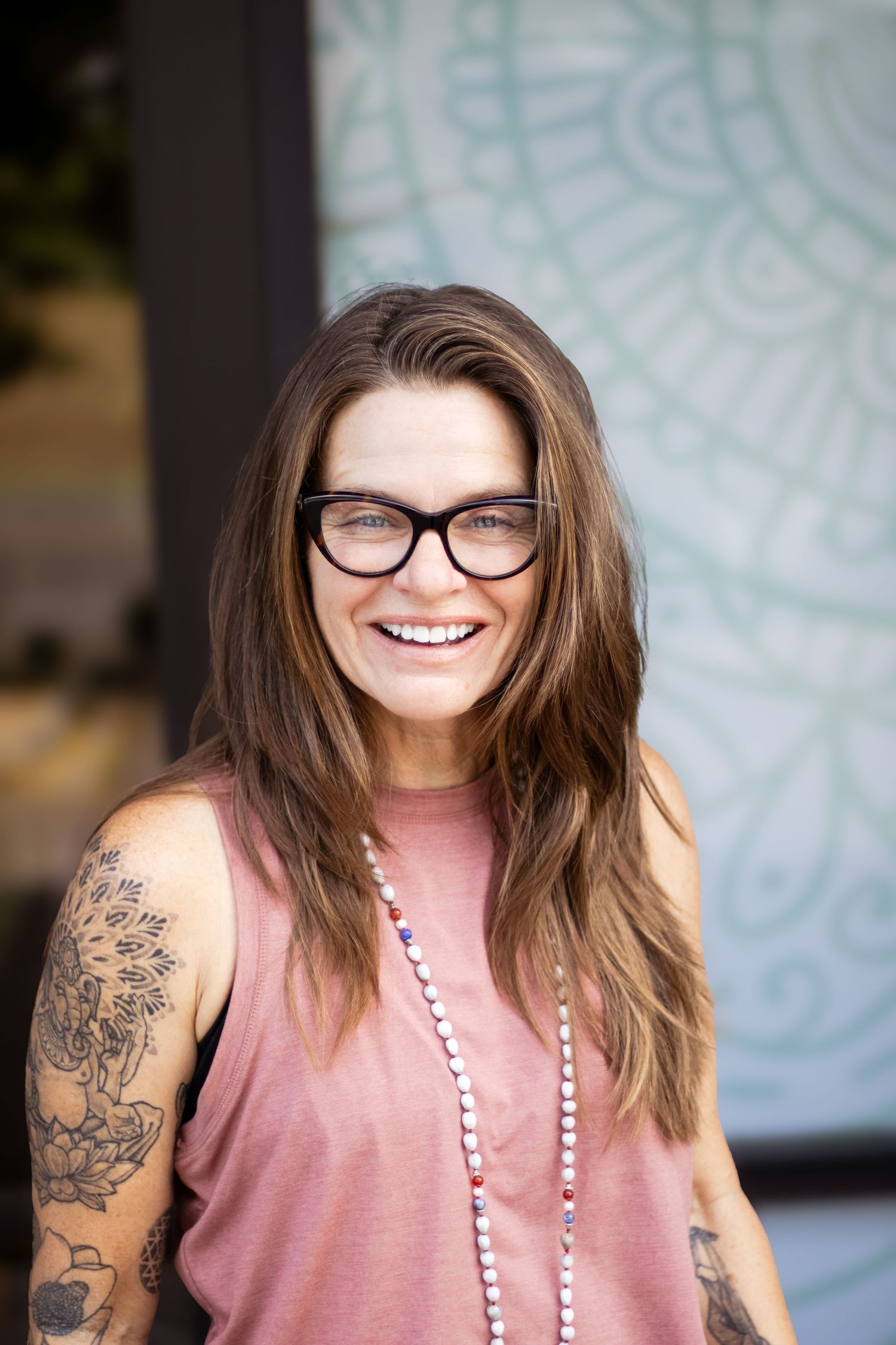 Casual portrait showing a beaded necklace, pink top and arm tattoo against a light patterned wall.