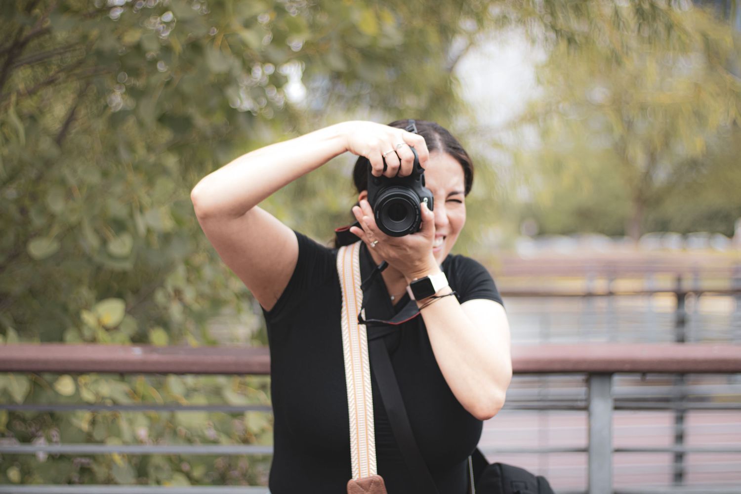 A photographer in black clothing takes photos outdoors in a natural setting with trees and sunlight.