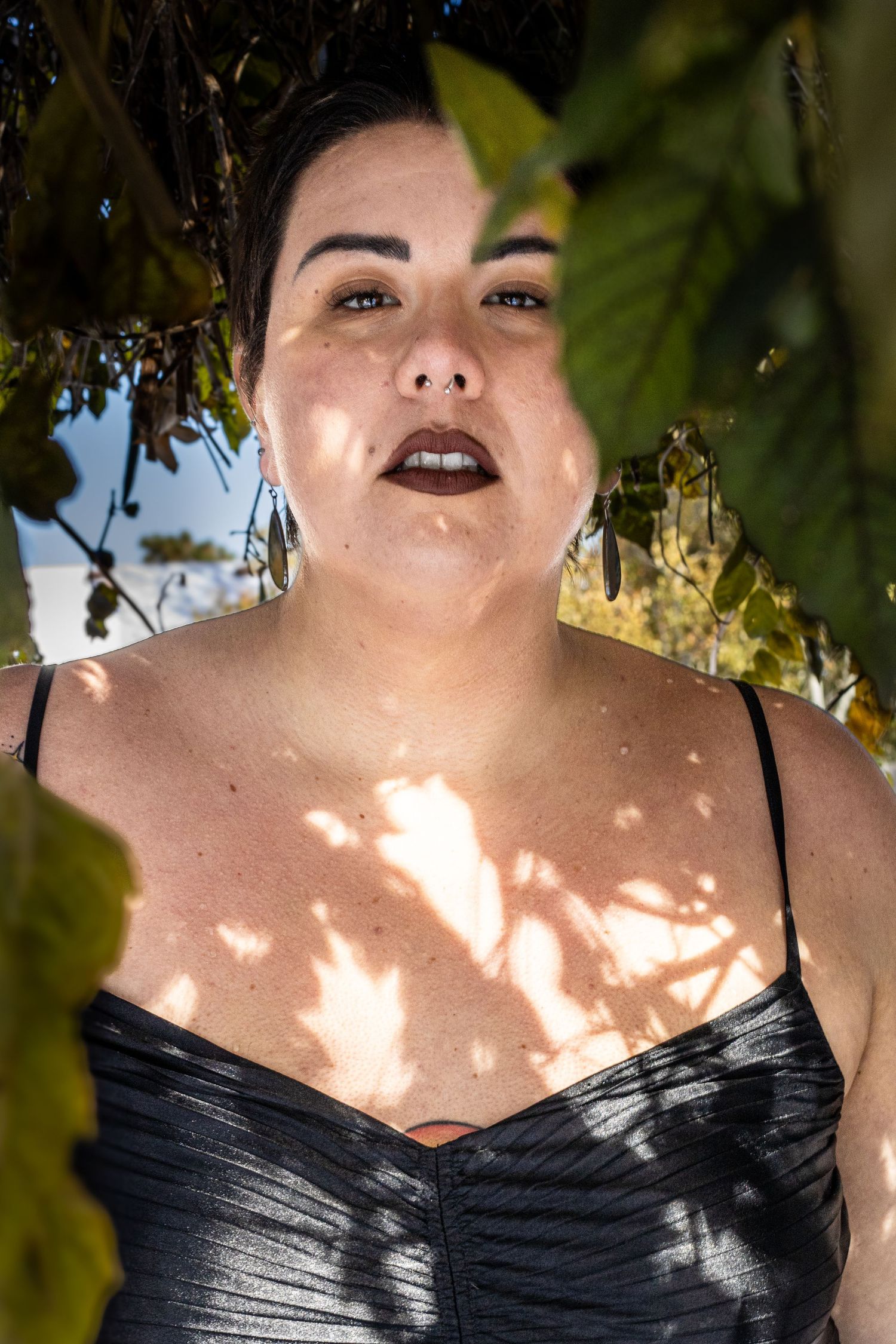 A person wearing dark lipstick and black clothing poses in dappled sunlight filtering through tree leaves.