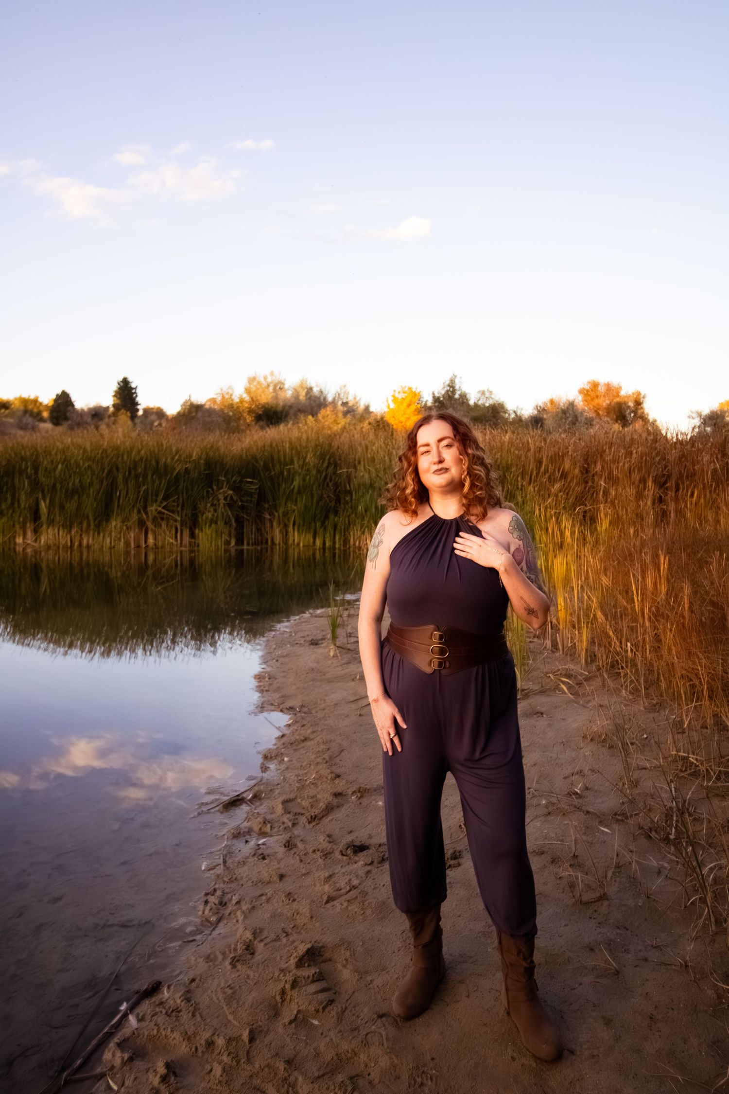 A figure in dark clothing stands on a sandy beach near tall reeds and water at sunset.