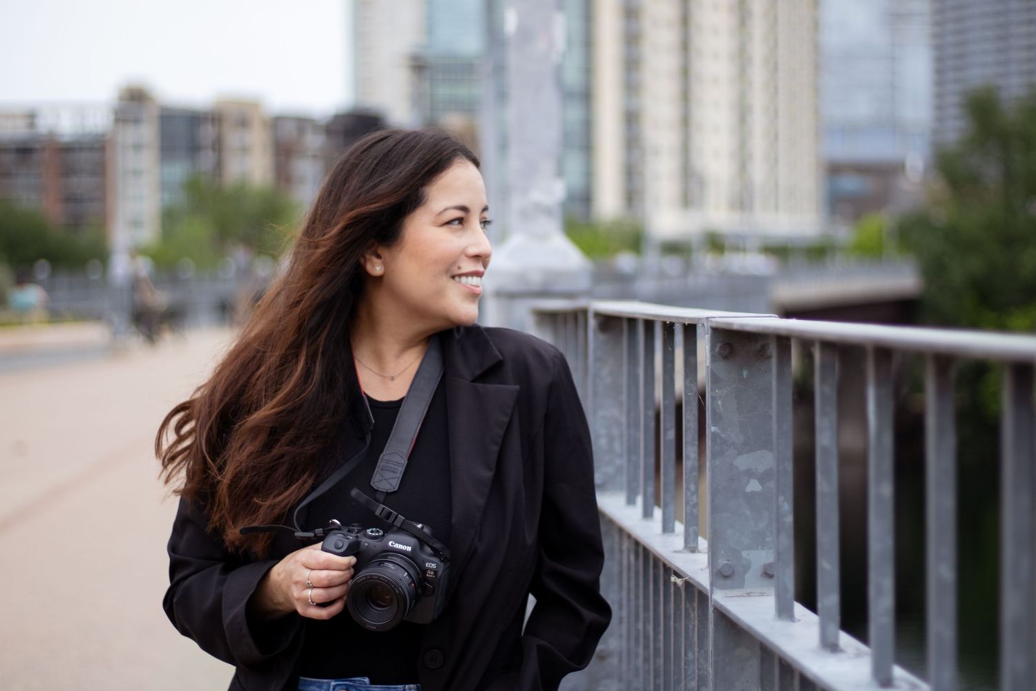 A photographer with long dark hair and a black jacket holds a camera while standing on an urban bridge.