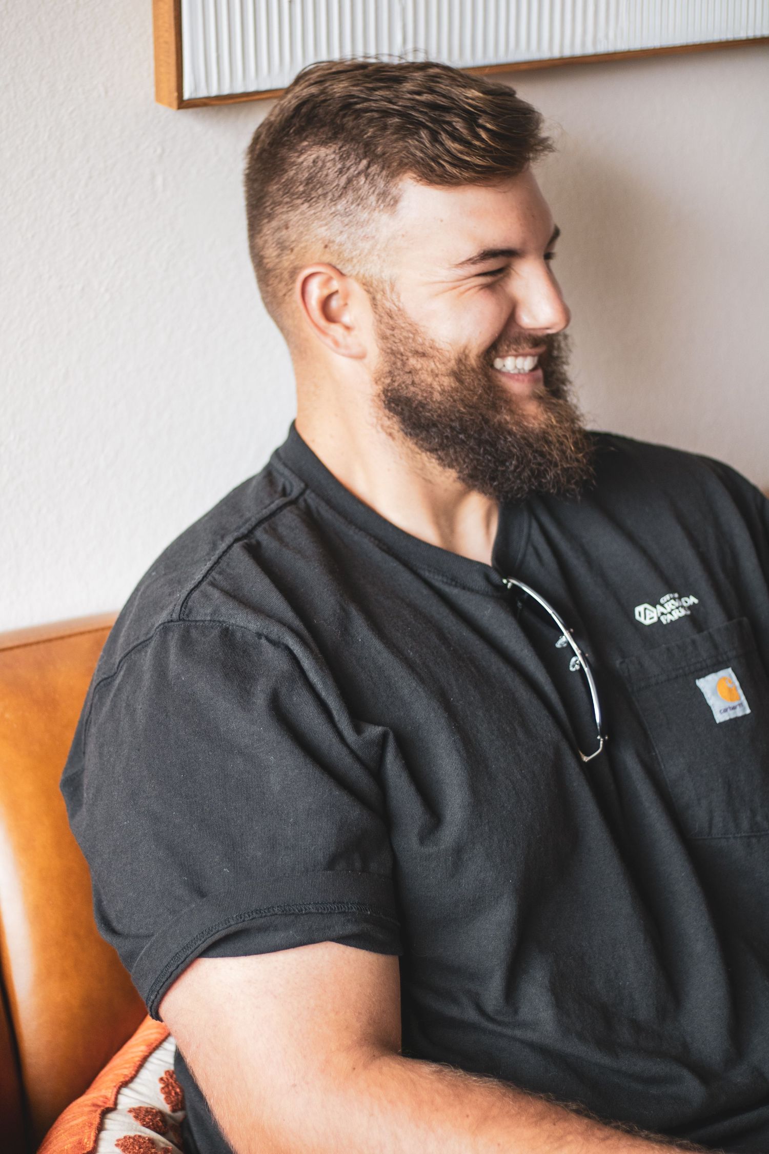 A bearded man in a black Carhartt t-shirt smiles while sitting on a leather chair.