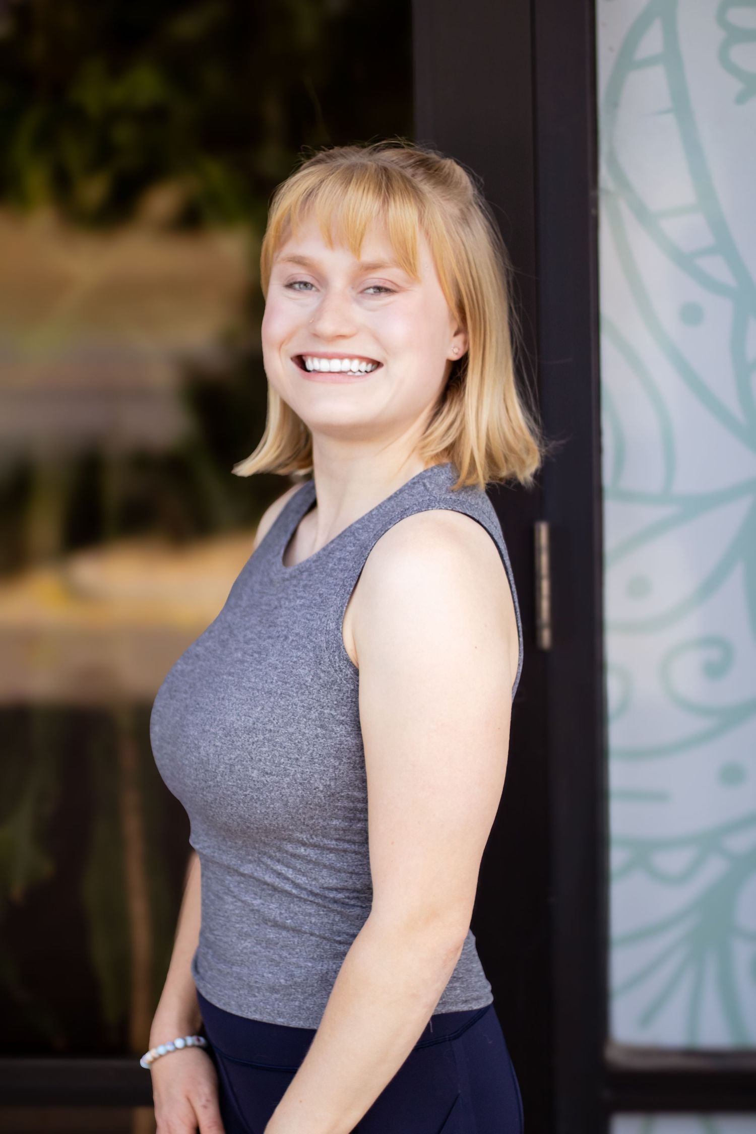 A person in a sleeveless grey top smiles warmly while standing in front of a doorway with decorative glass panels.