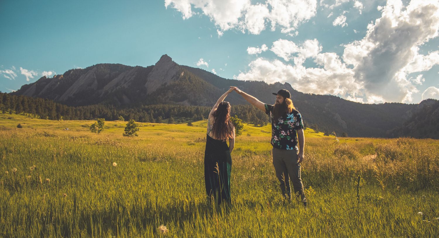 Two people hiking through a golden meadow with majestic mountain peaks and blue skies in the background.