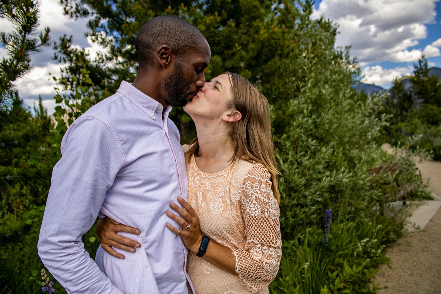 A couple shares a romantic kiss on a sunny day in a lush green outdoor setting.