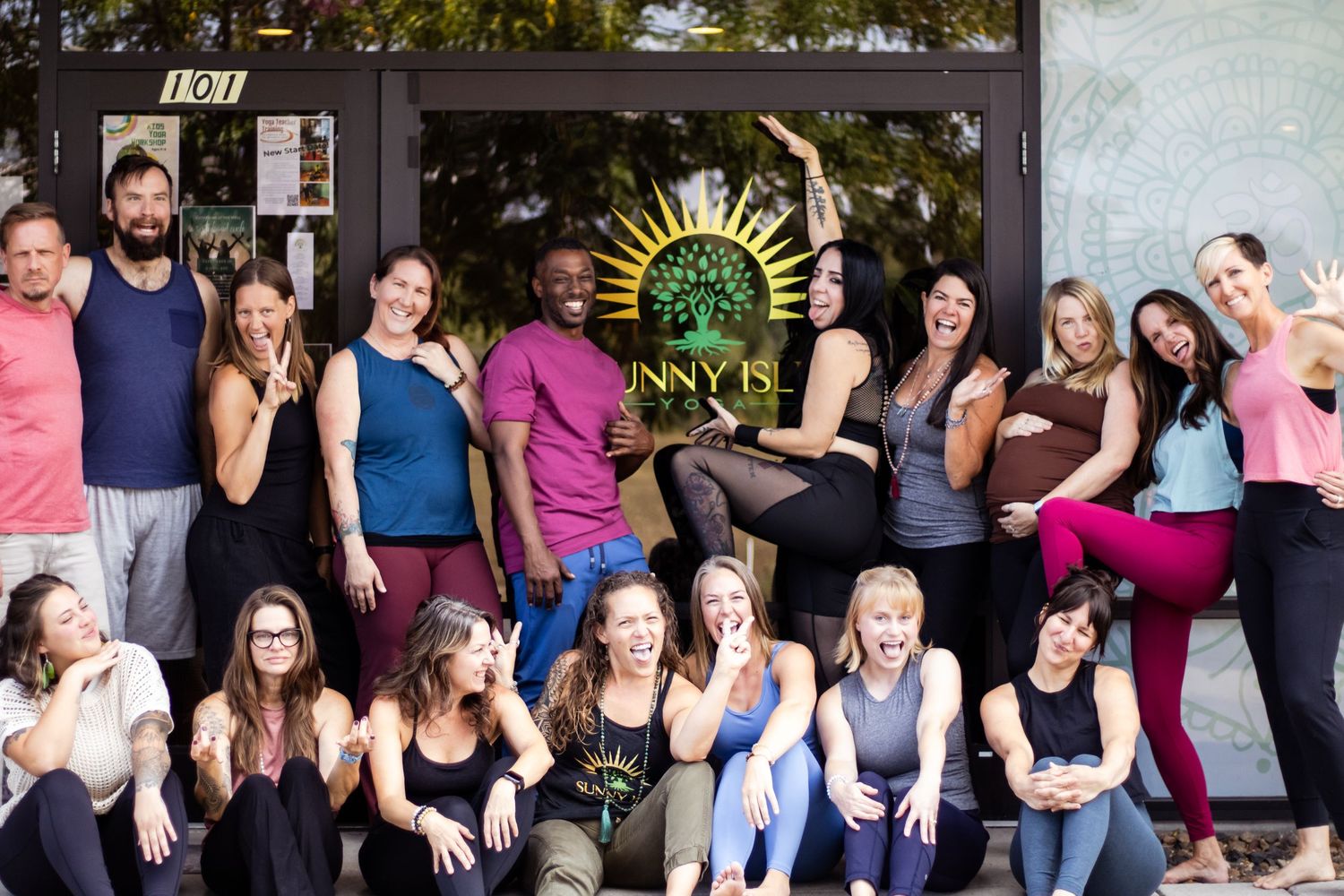 A group of yoga practitioners pose together in athletic wear outside against a wall with a decorative sun artwork.