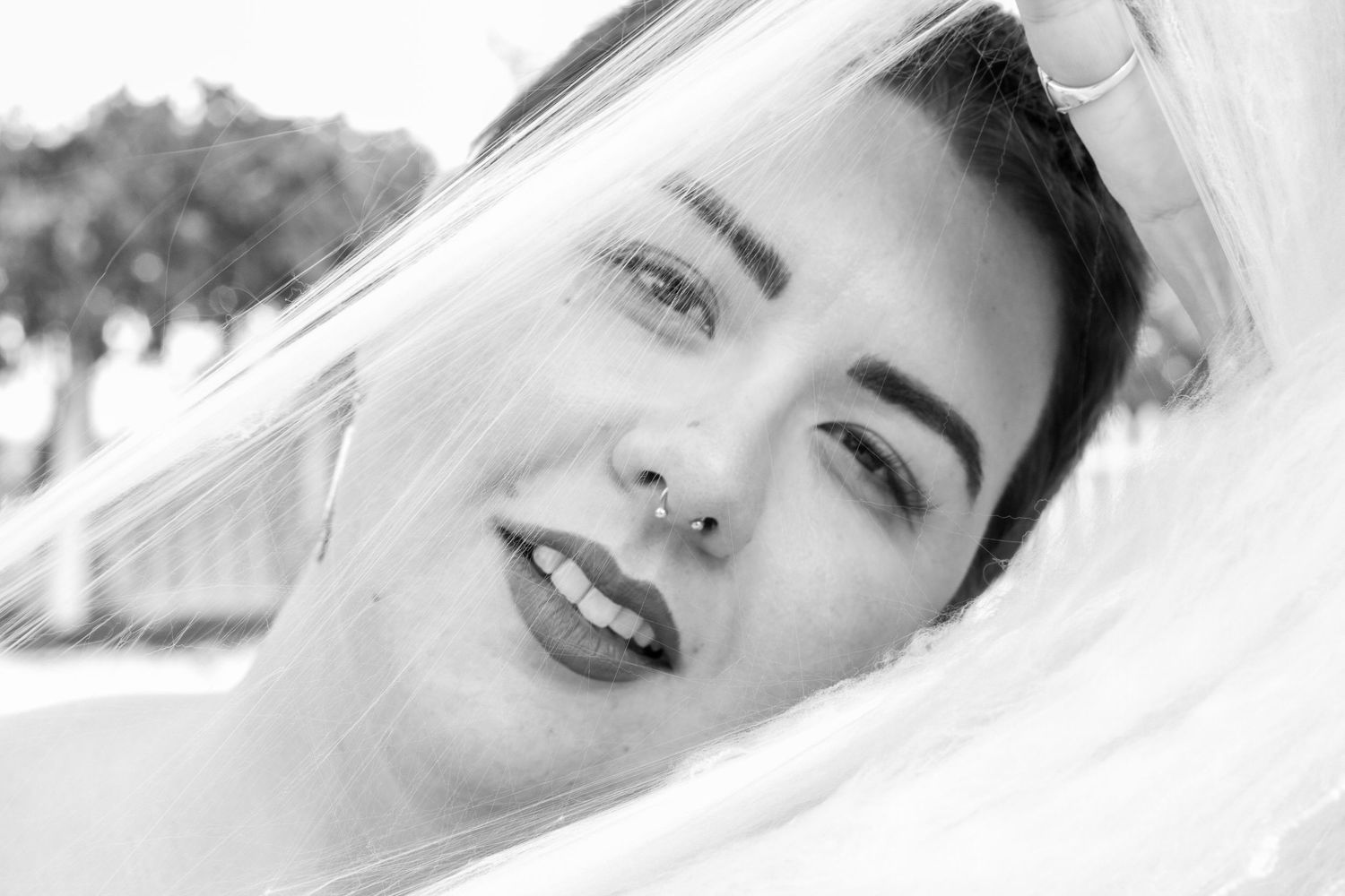 Black and white close up portrait of a smiling person with dark lipstick and nose piercing under a white veil.