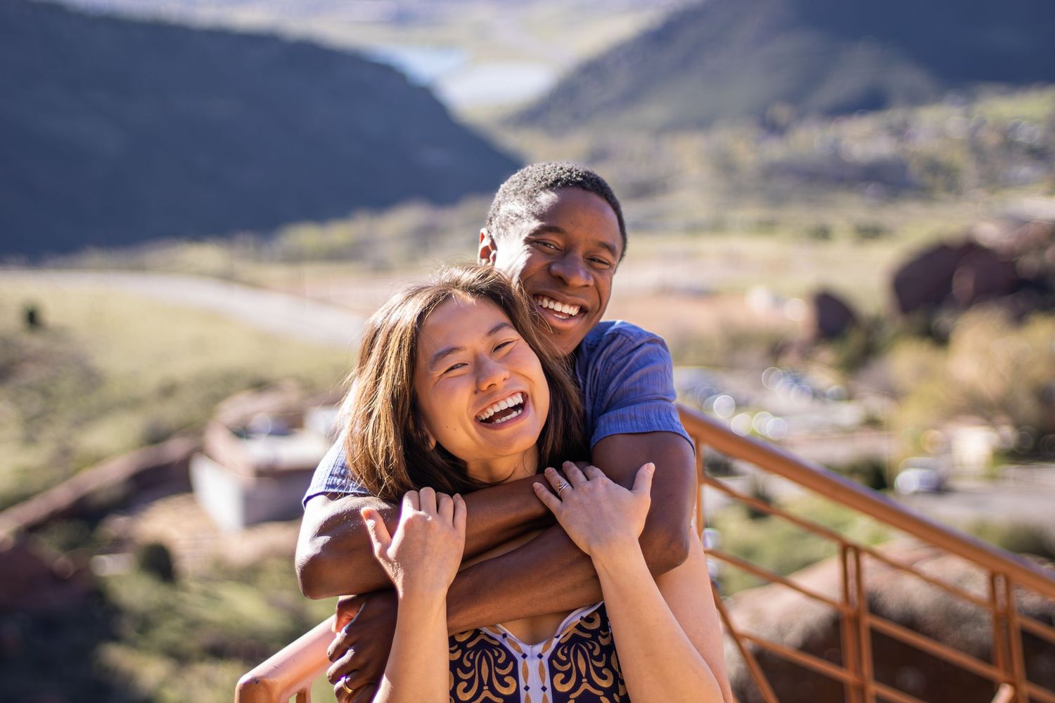 Two friends share a joyful embrace while enjoying a scenic mountain landscape on a sunny day.