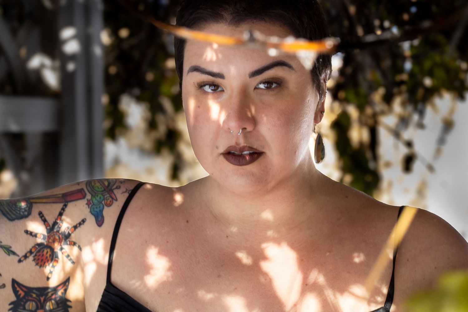 A woman in a black dress and dark lipstick poses outdoors with dappled sunlight falling across her face.