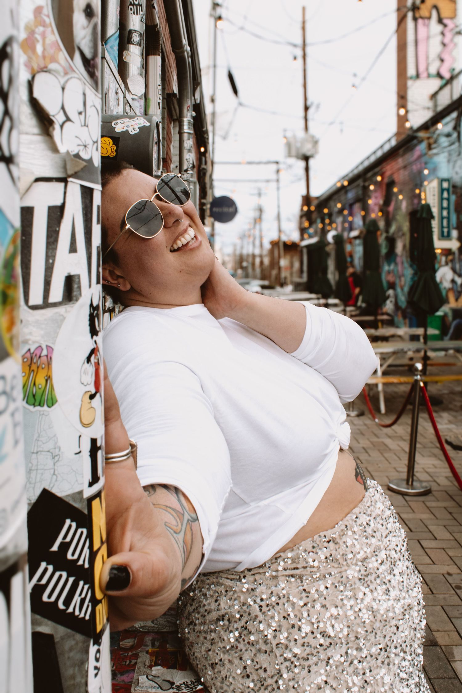 Woman in white t-shirt and sequined skirt poses playfully against white wall in urban setting.