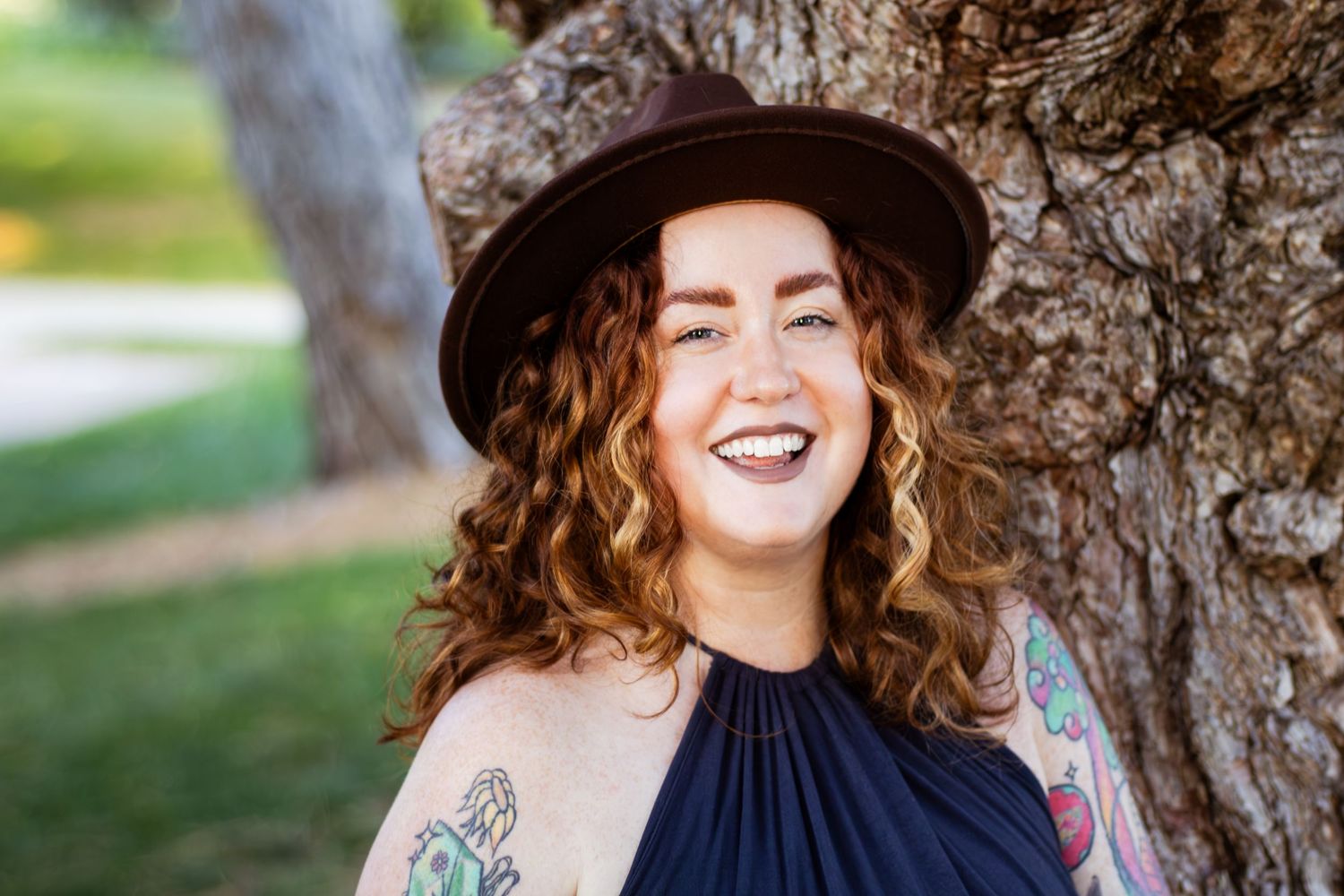 A joyful person in a black hat smiles while standing next to a large tree trunk in an outdoor setting.
