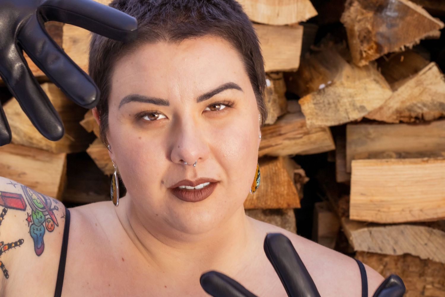 Person wearing black gloves poses in front of stacked firewood with dark lipstick and earrings.