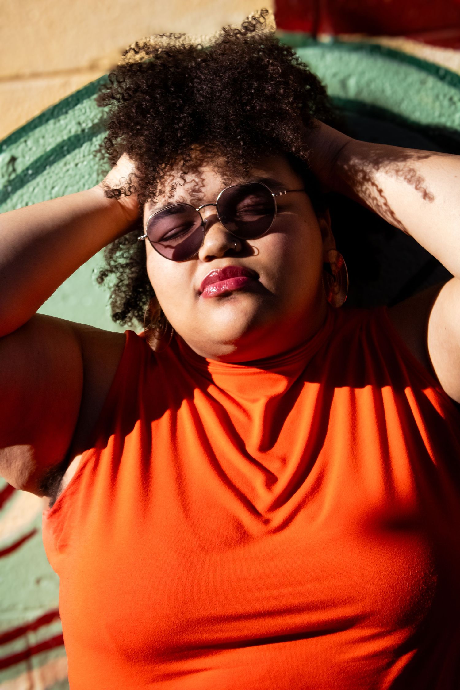 A person with curly hair wearing a bright orange turtleneck sweater poses against a turquoise wall.