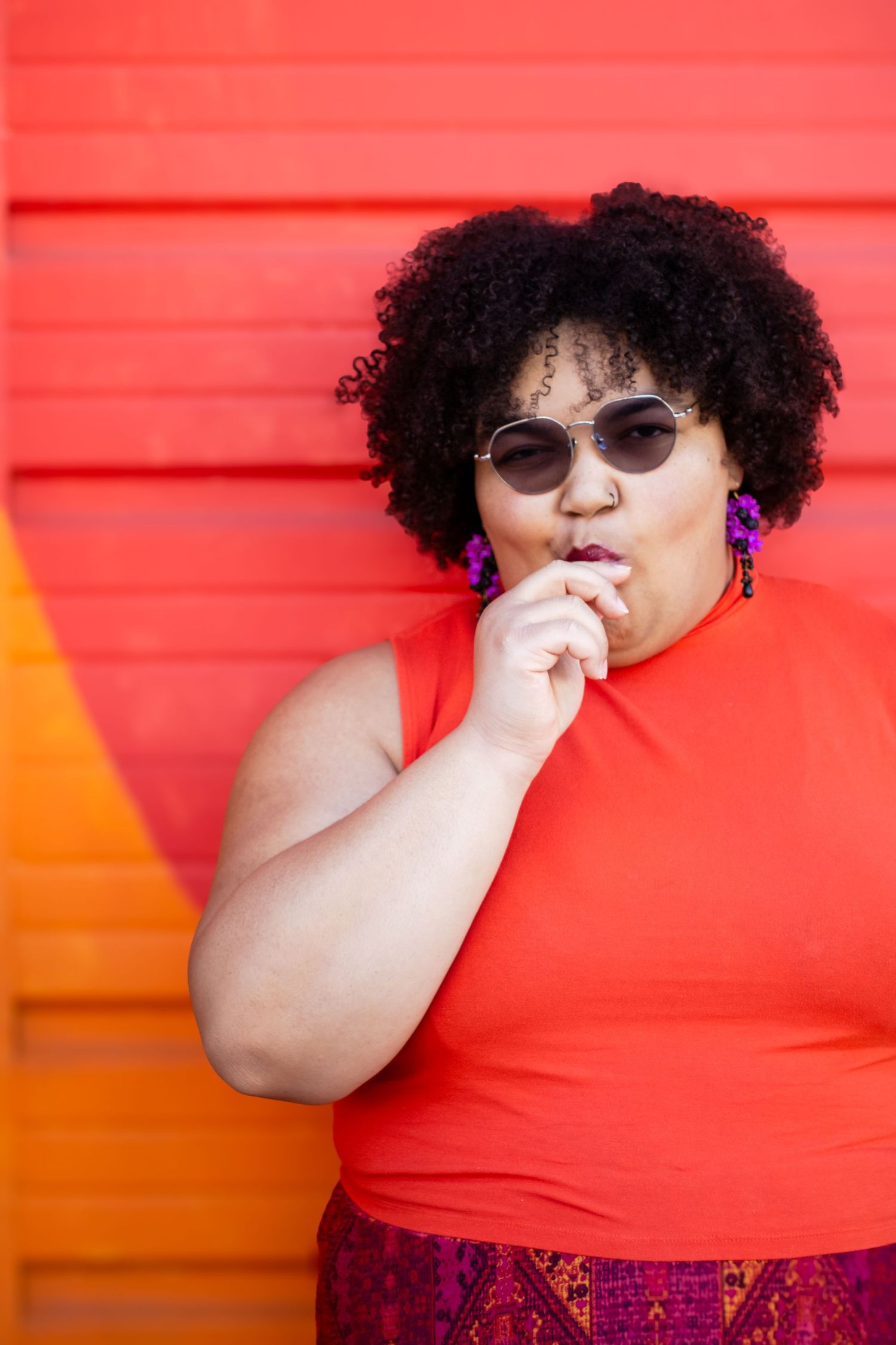 A person in a red sleeveless top and sunglasses poses against a vibrant orange and red wall background.